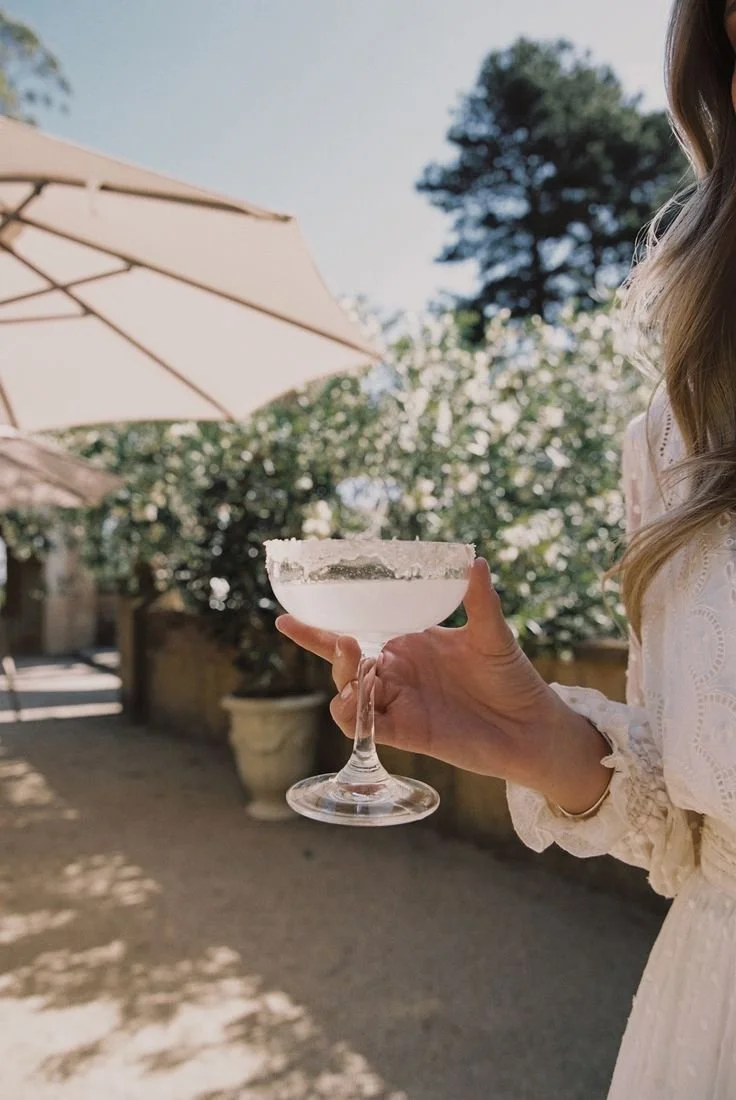 Person holding a cocktail glass with a salted rim outdoors on a sunny day, with a patio umbrella and greenery in the background.