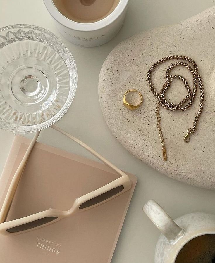 A flat lay of a glass of water, a cup of coffee, a jewelry plate with a gold ring and a chain necklace, a pink tray with sunglasses, and a white ceramic mug with black coffee.