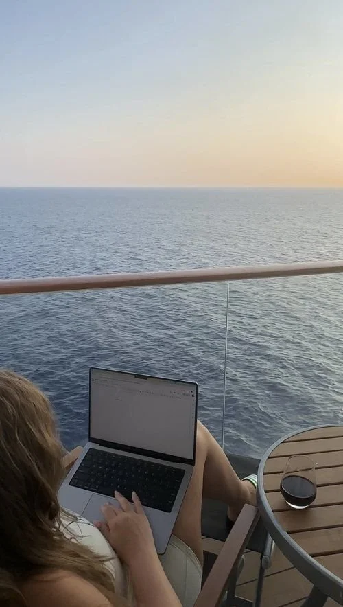 Person with long hair sitting on a cruise ship balcony, working on a laptop, with a glass of red wine on a round wooden table, overlooking the ocean during sunset.