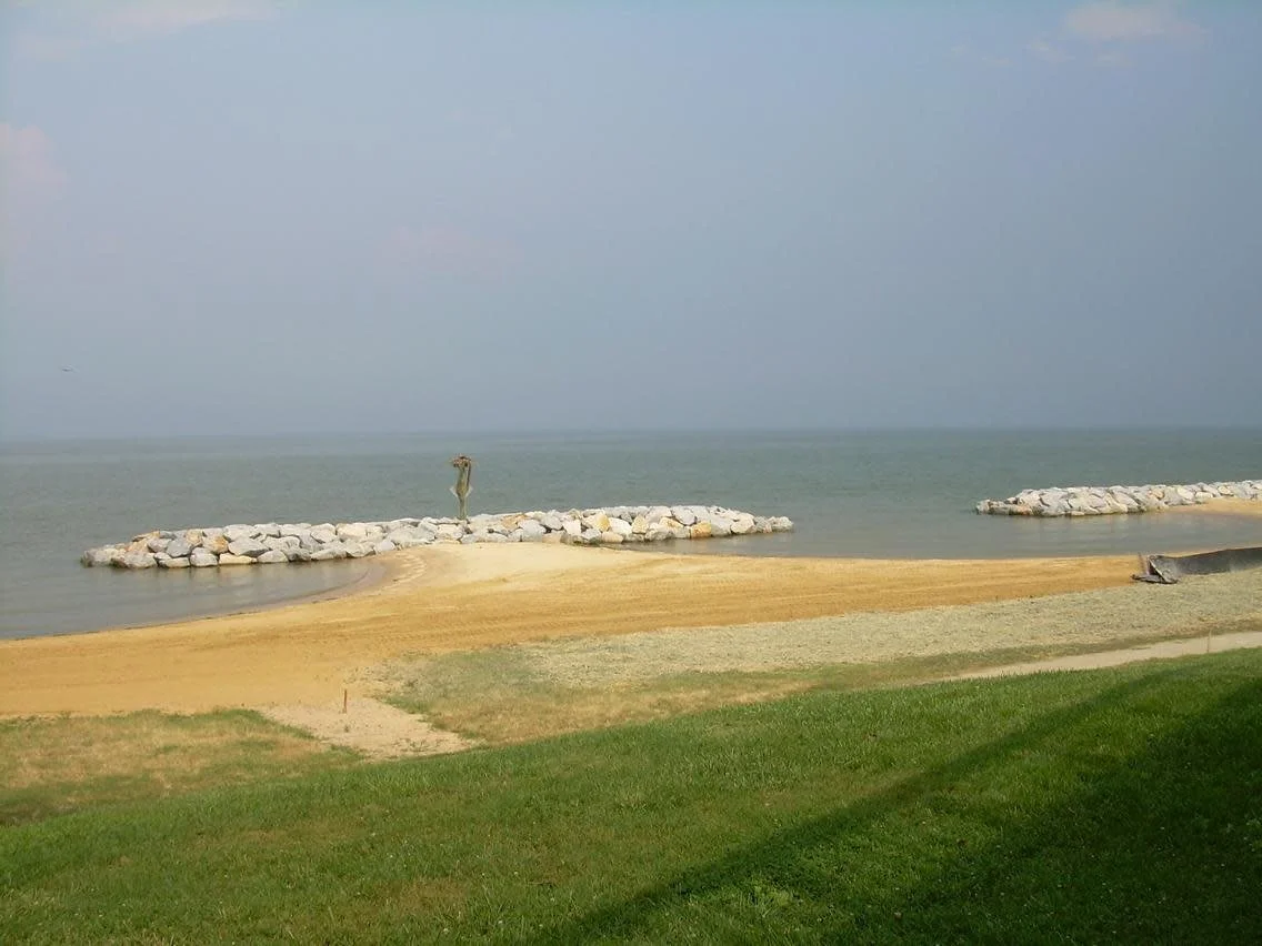 Beach scene with grassy area in the foreground, sandy shoreline, breakwater with rocks, and calm water beyond, overcast sky.