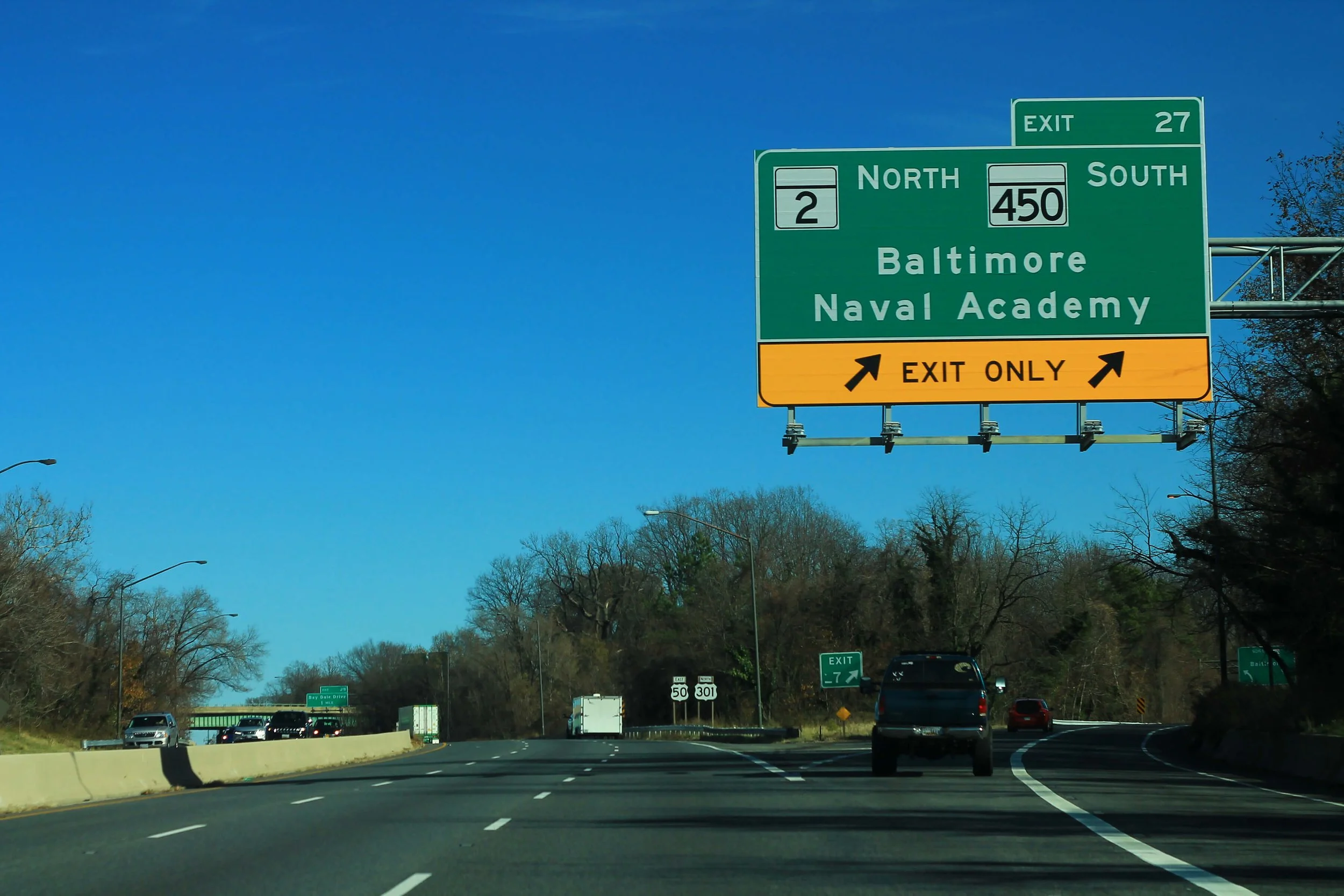 Highway sign indicating the exit for Baltimore Naval Academy and routes 2, 450, and 27, with the sky and trees in the background.