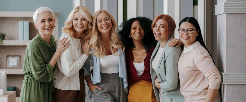 Six women smiling and standing together in a modern office setting, embracing each other's shoulders.