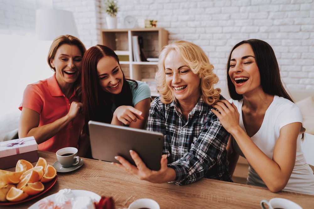 Four women laughing and sharing a tablet at a table with oranges, tea, and gifts in a cozy, well-lit room.