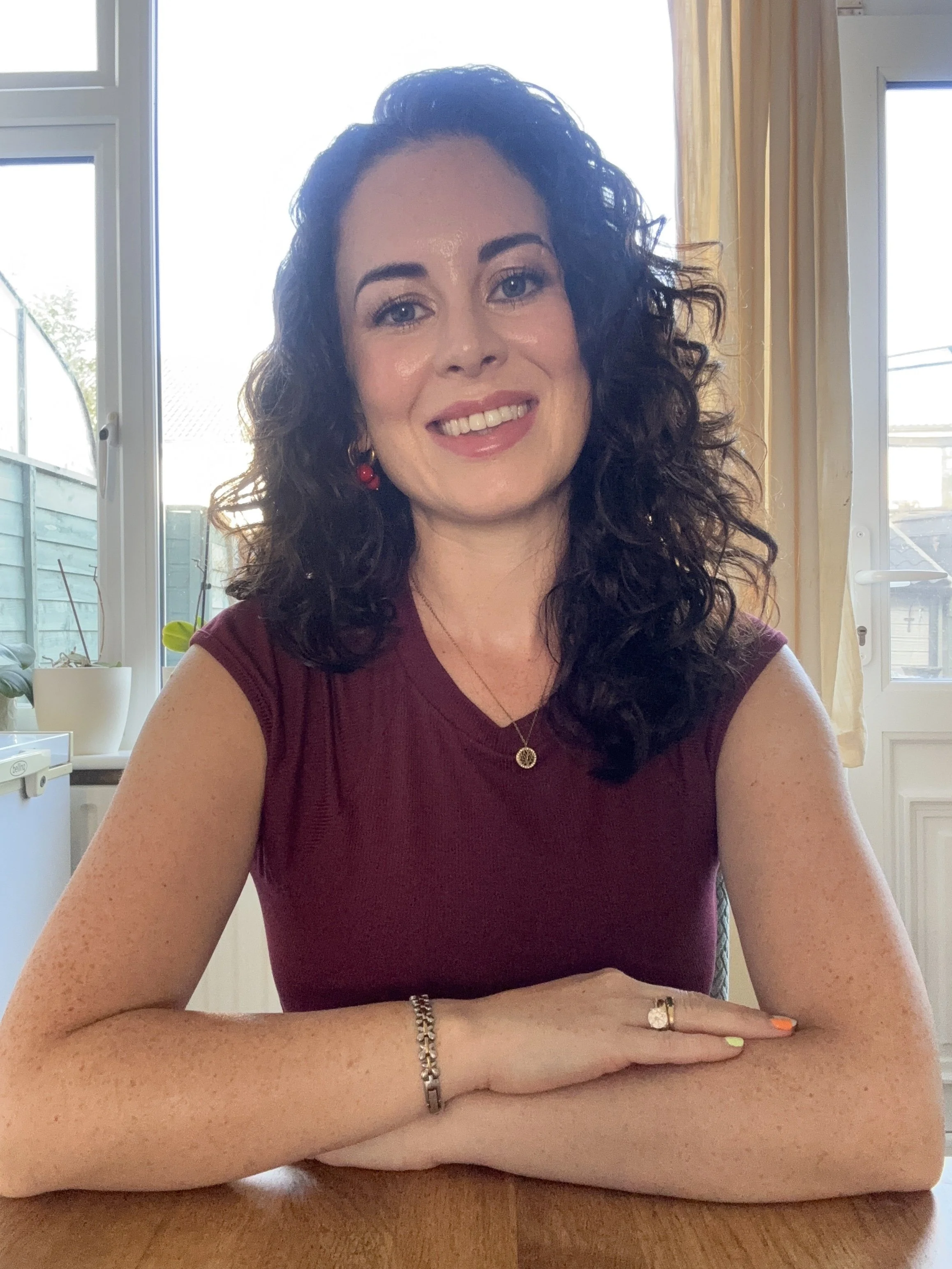 A woman with curly dark hair, light skin, wearing a maroon sleeveless top, gold jewelry including earrings, a necklace, and rings, is sitting at a wooden table inside a room with large windows and yellow curtains, smiling at the camera.