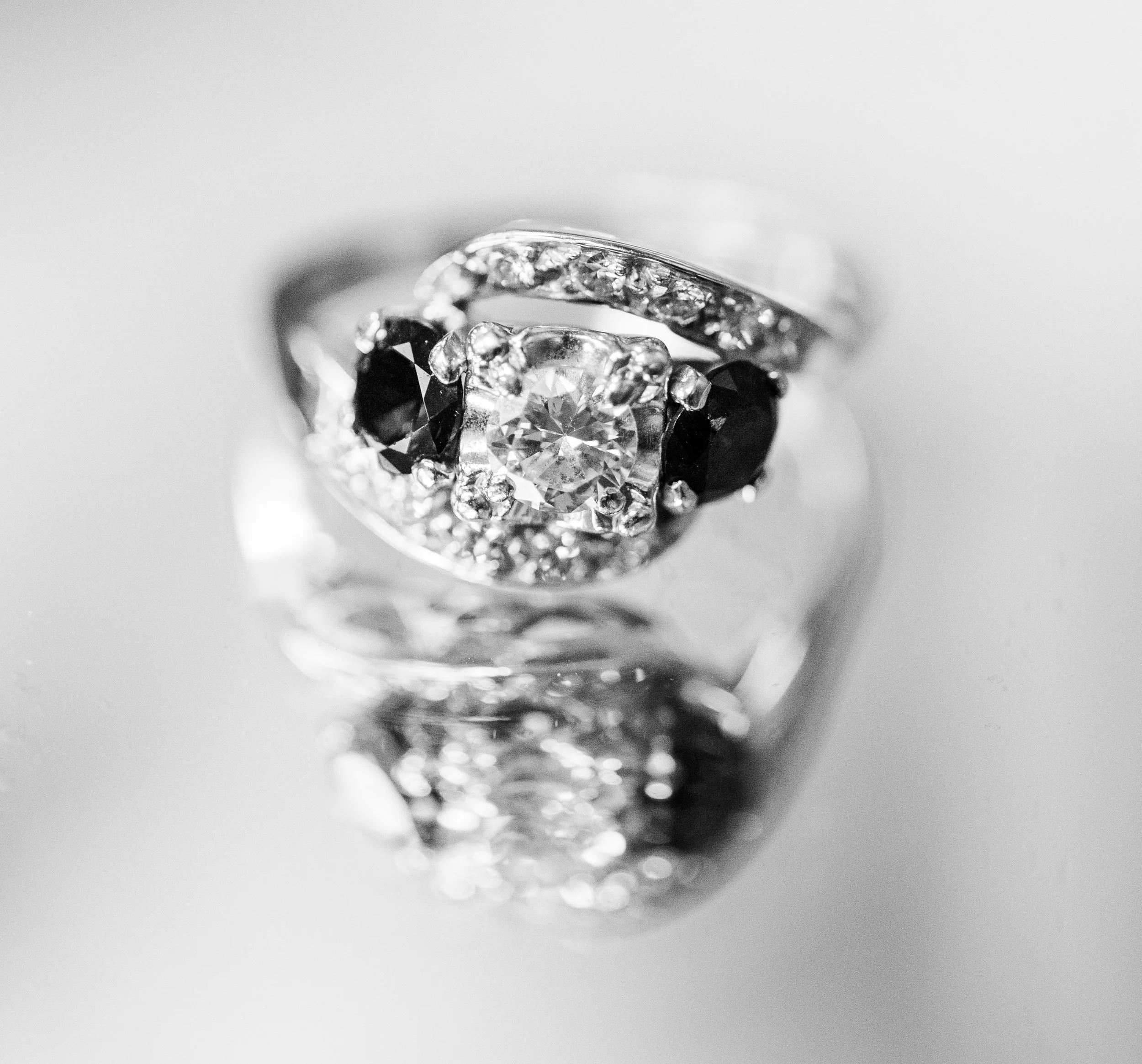 Close-up view of a silver ring with a large central diamond, accented by smaller black and white diamonds, and a reflection on a smooth surface.
