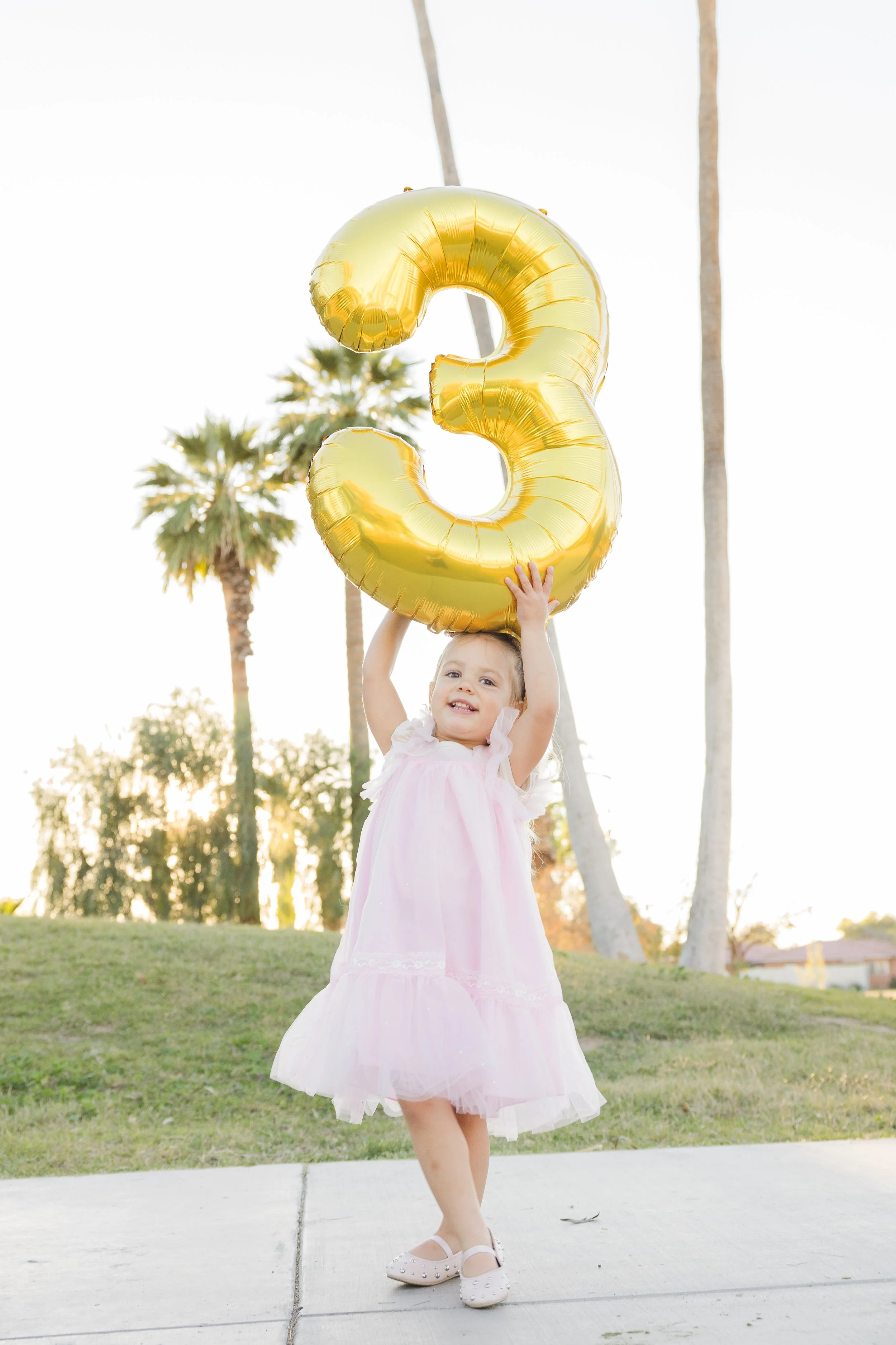 A young girl in a pink dress holding a large gold balloon shaped like the number three outdoors during sunset, with palm trees in the background.