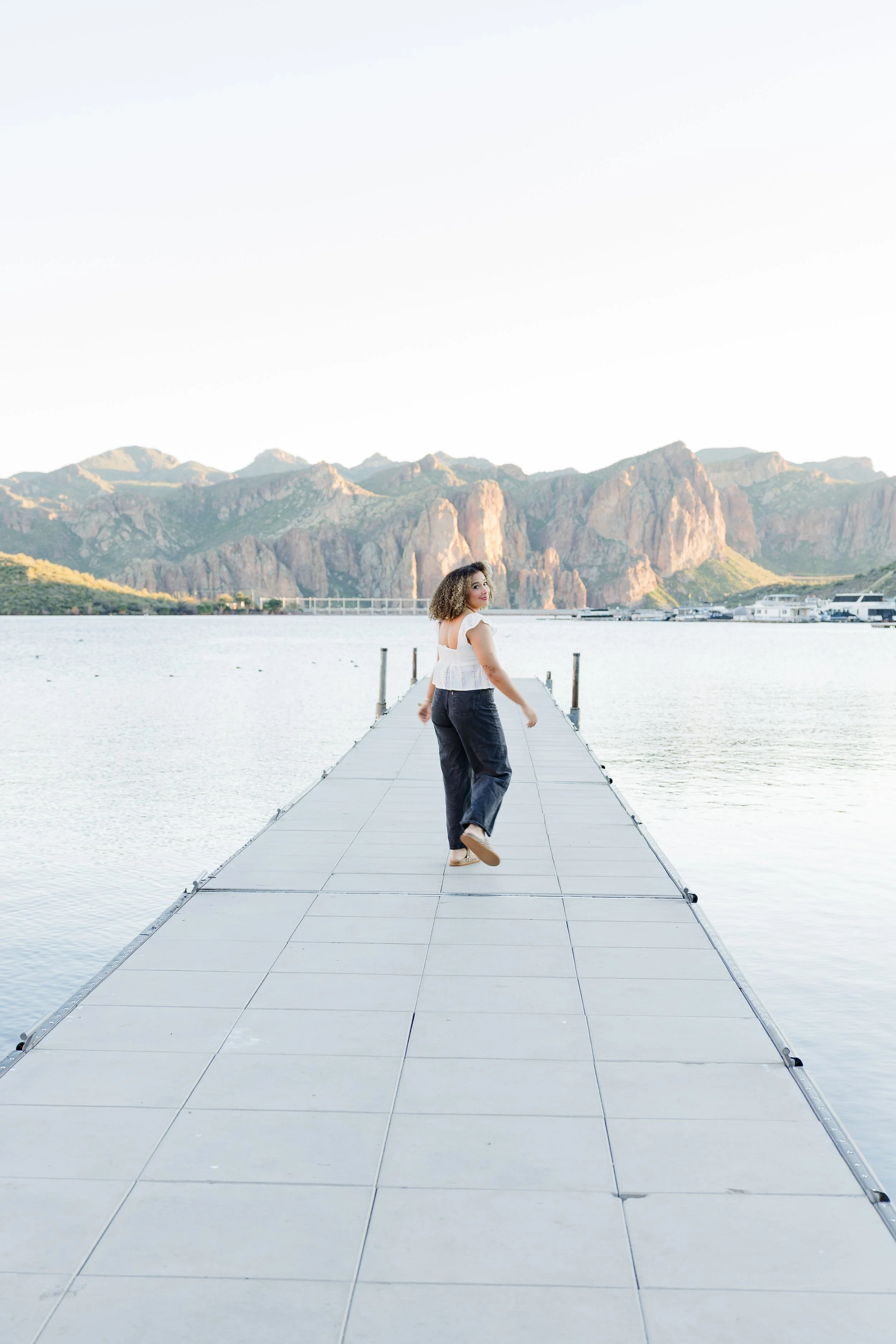 A young woman with long wavy blonde hair smiling, wearing a maroon graduation gown and white stole with Greek letters, standing on a grassy field with trees and a building in the background.
