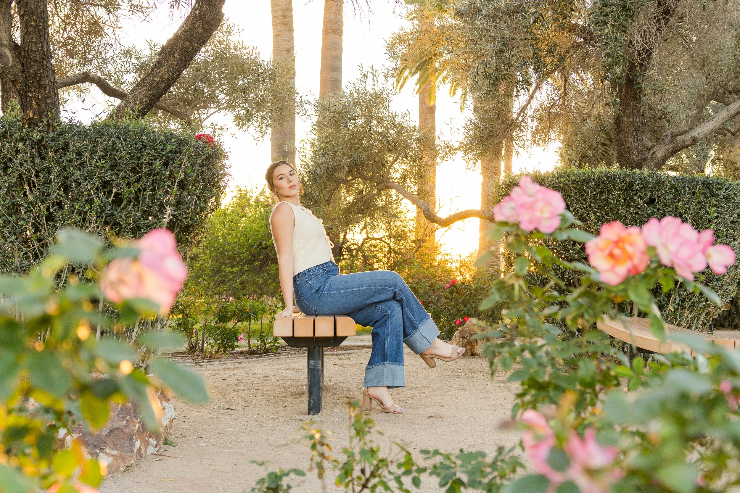 A woman sitting on a park bench surrounded by pink flowers and lush greenery during sunset.