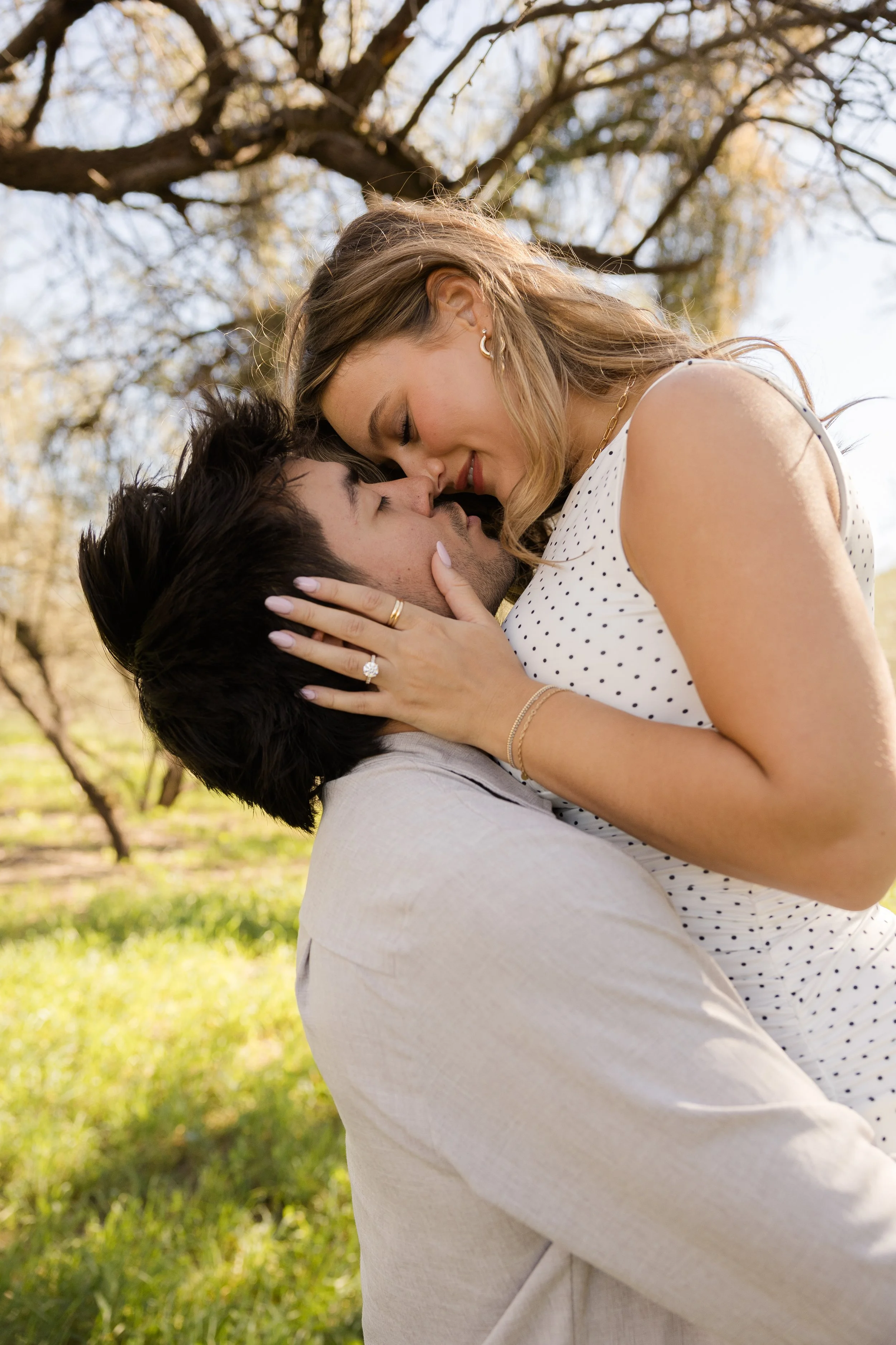 A couple sharing a tender moment outdoors, with the woman holding the man's face as they lean in close, their noses touching, under a tree during daylight.