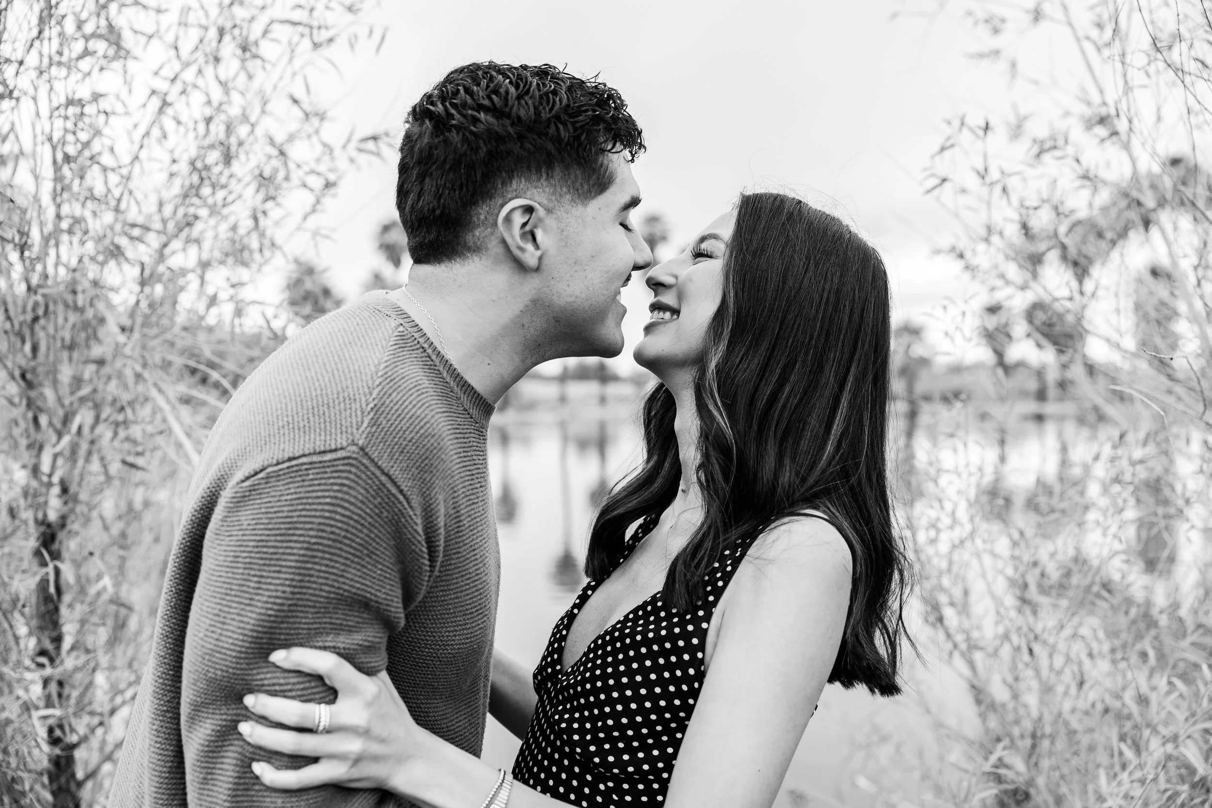 A black and white photo of a couple smiling and about to kiss outdoors, surrounded by trees.