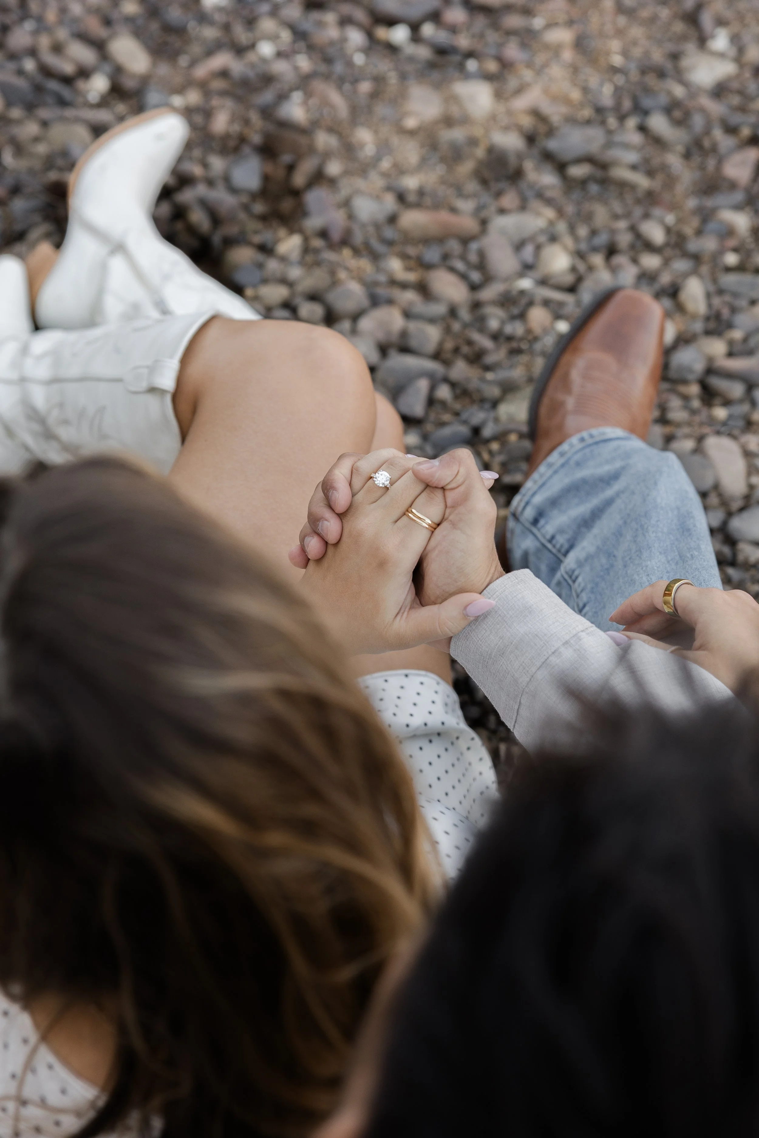 A couple holding hands, sitting on rocky ground, with one person wearing white boots and the other wearing brown shoes. The woman has a diamond ring on her finger.
