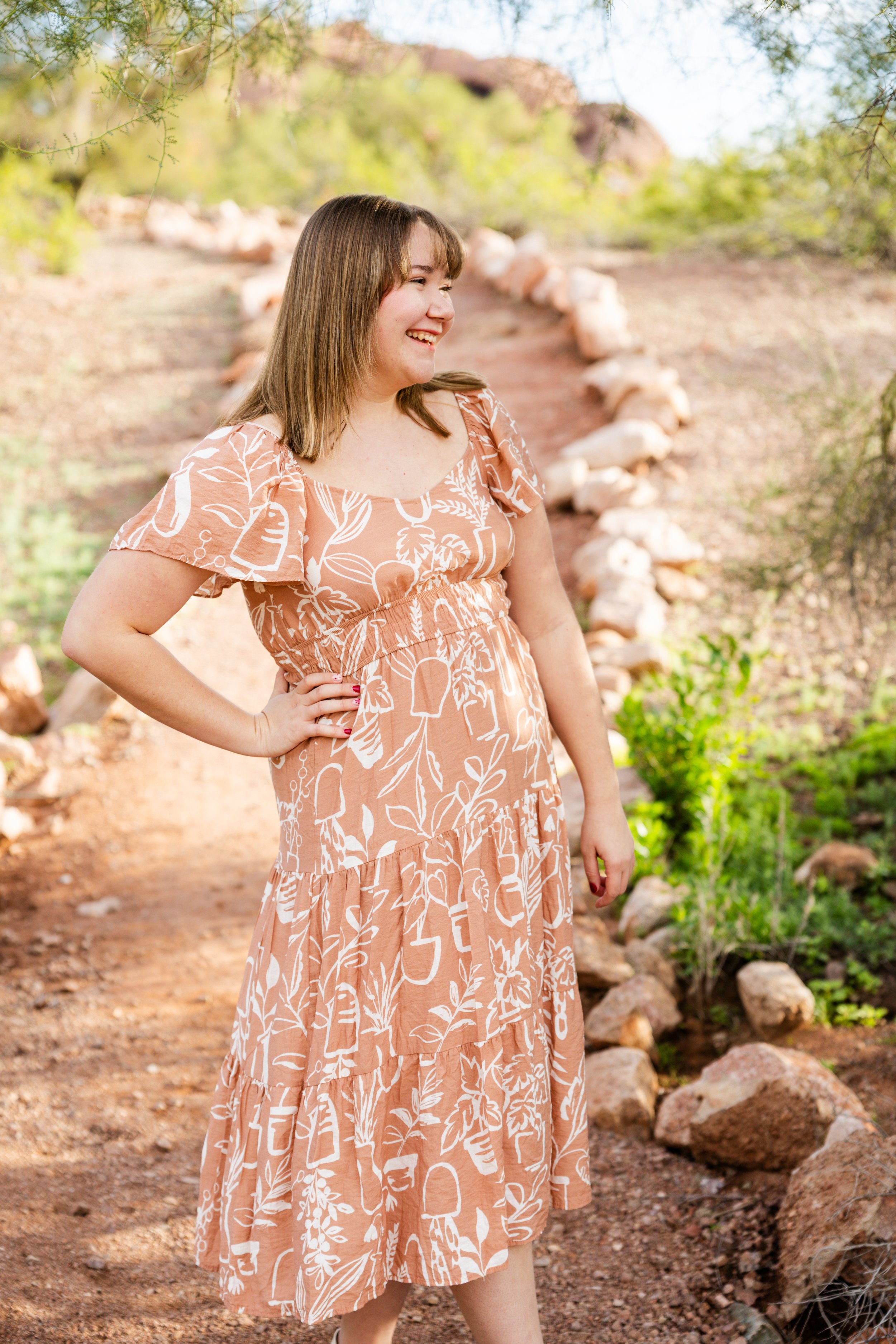 A young woman with shoulder-length brown hair smiling and standing outdoors on a rocky dirt path surrounded by desert vegetation, rocks, and small green plants.