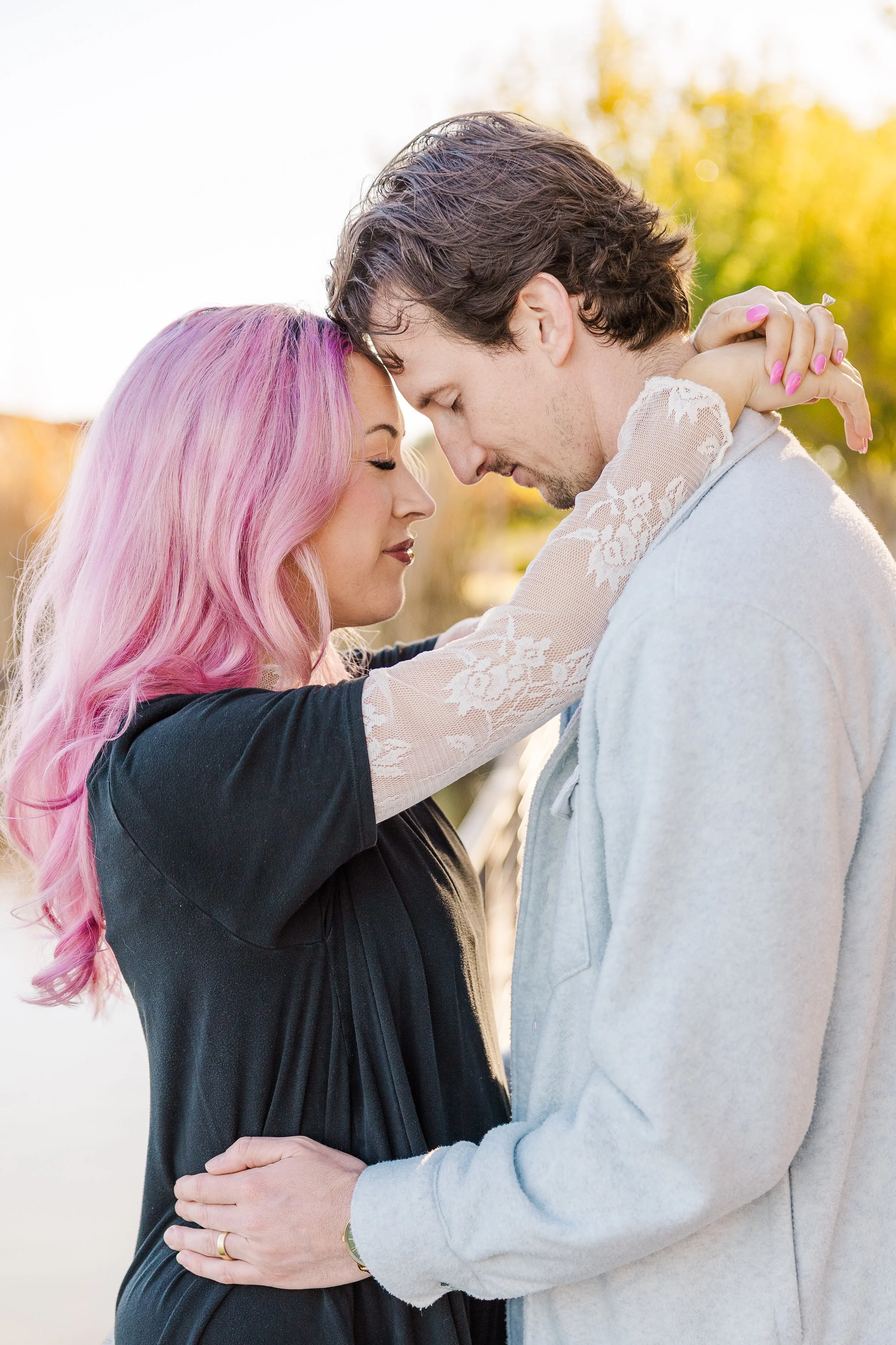 A couple standing close with foreheads touching, outdoors during sunset, with trees in the background.