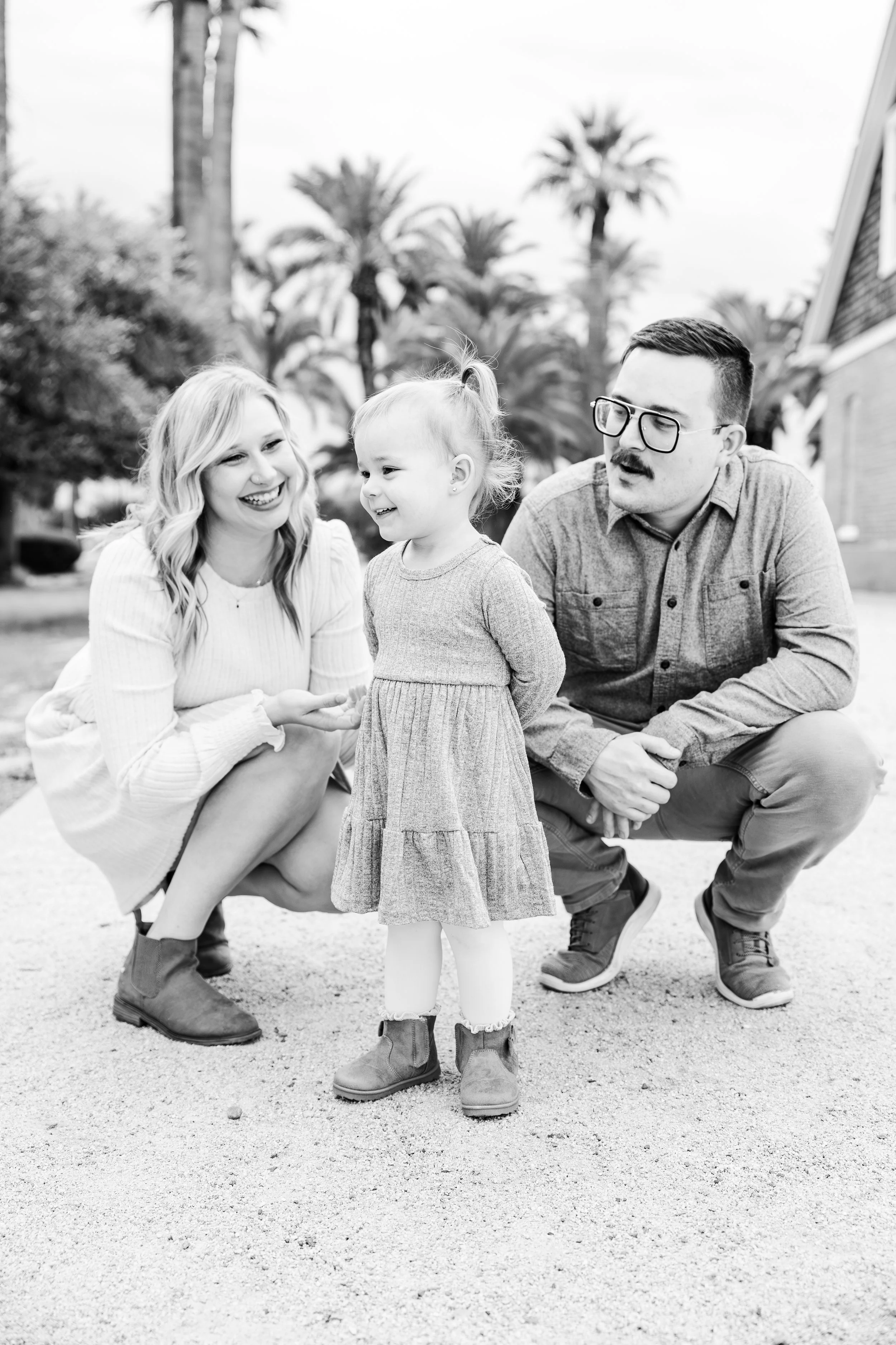 A black-and-white photo of a family outdoors with palm trees in the background. A woman and a man are crouching on either side of a young girl, all smiling and interacting.