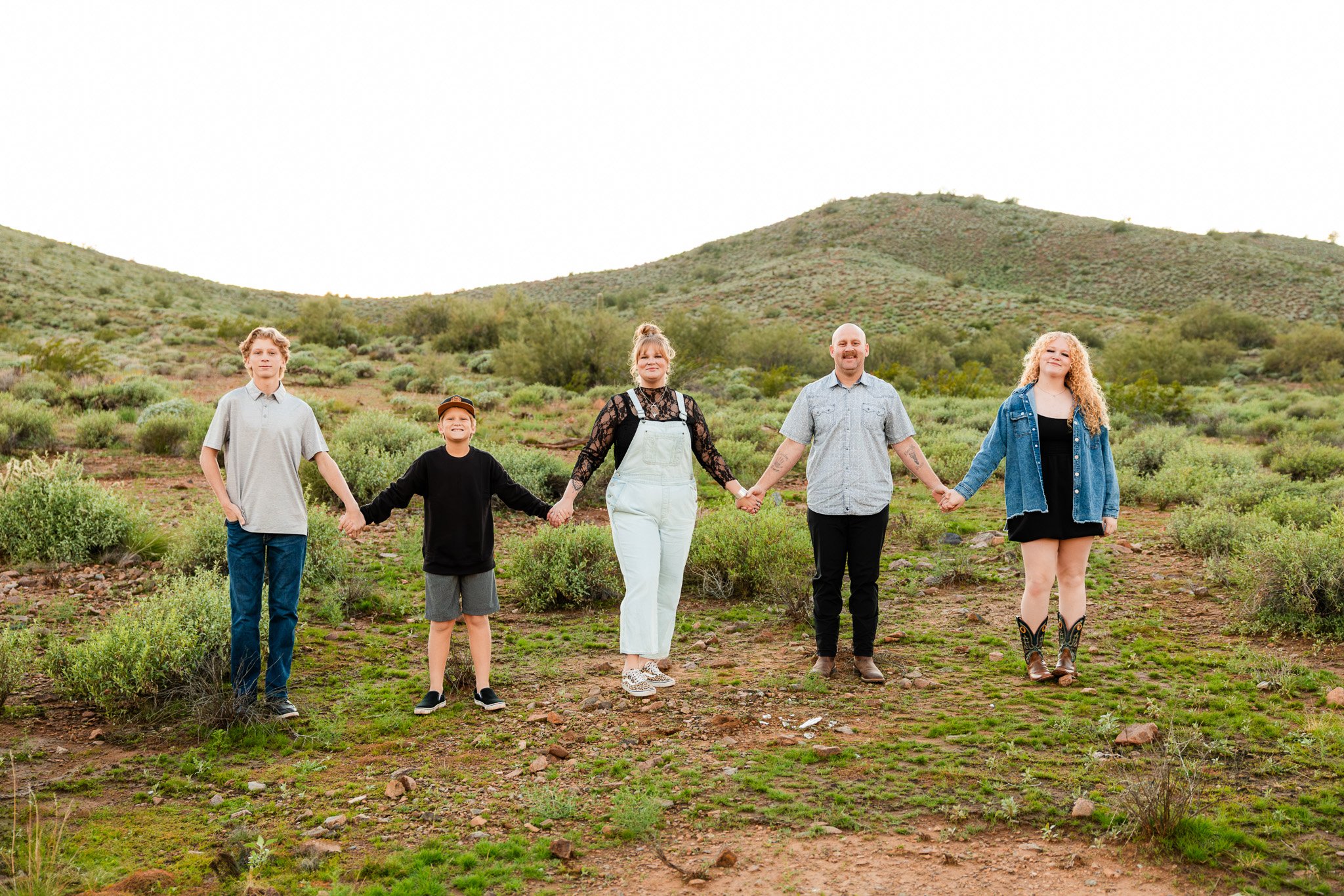 A happy family of three outdoors on a sunny day: a man, a woman, and a young girl. The man is wearing a white shirt and teal shorts, the woman is in a white dress, and the girl is in a pink top and white skirt. They are standing on a grassy path surrounded by trees and greenery.