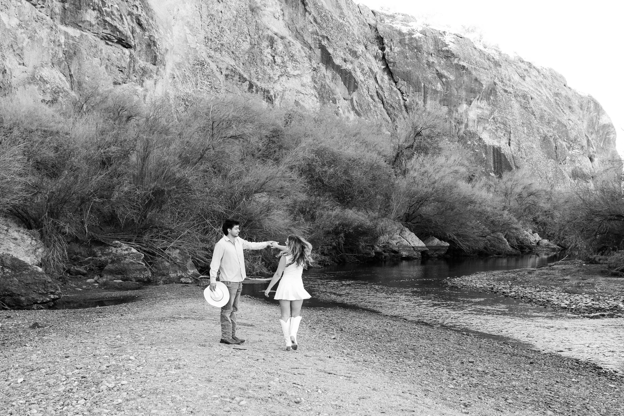 A black and white photo of a young man and woman on a riverbank, the man holding a hat and touching the woman's hand, with a rocky cliff and leafless trees in the background.