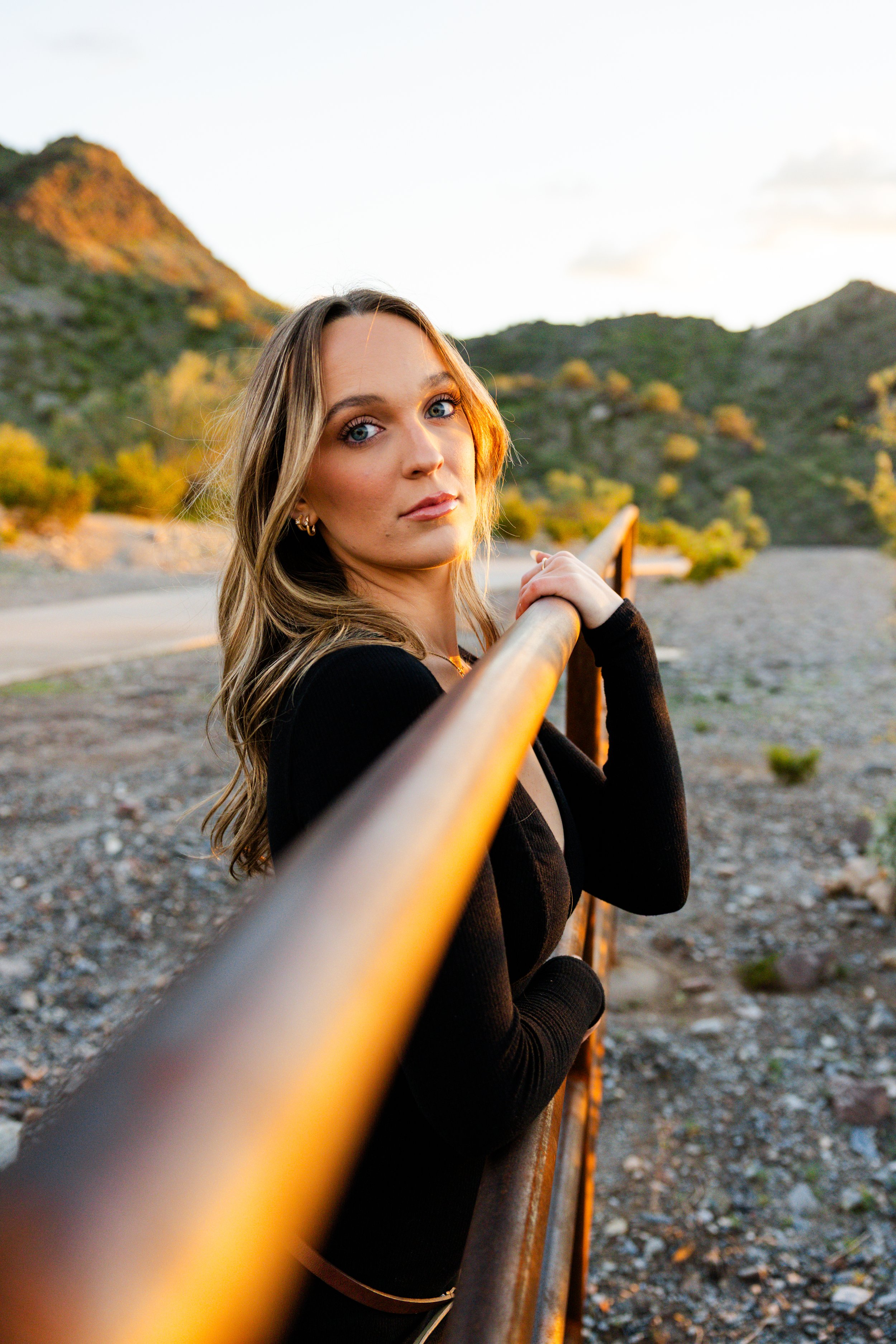 A woman with wavy, blonde hair wearing a black top is standing by a yellow railing on a bridge or walkway, with mountainous terrain and green trees in the background during sunset.