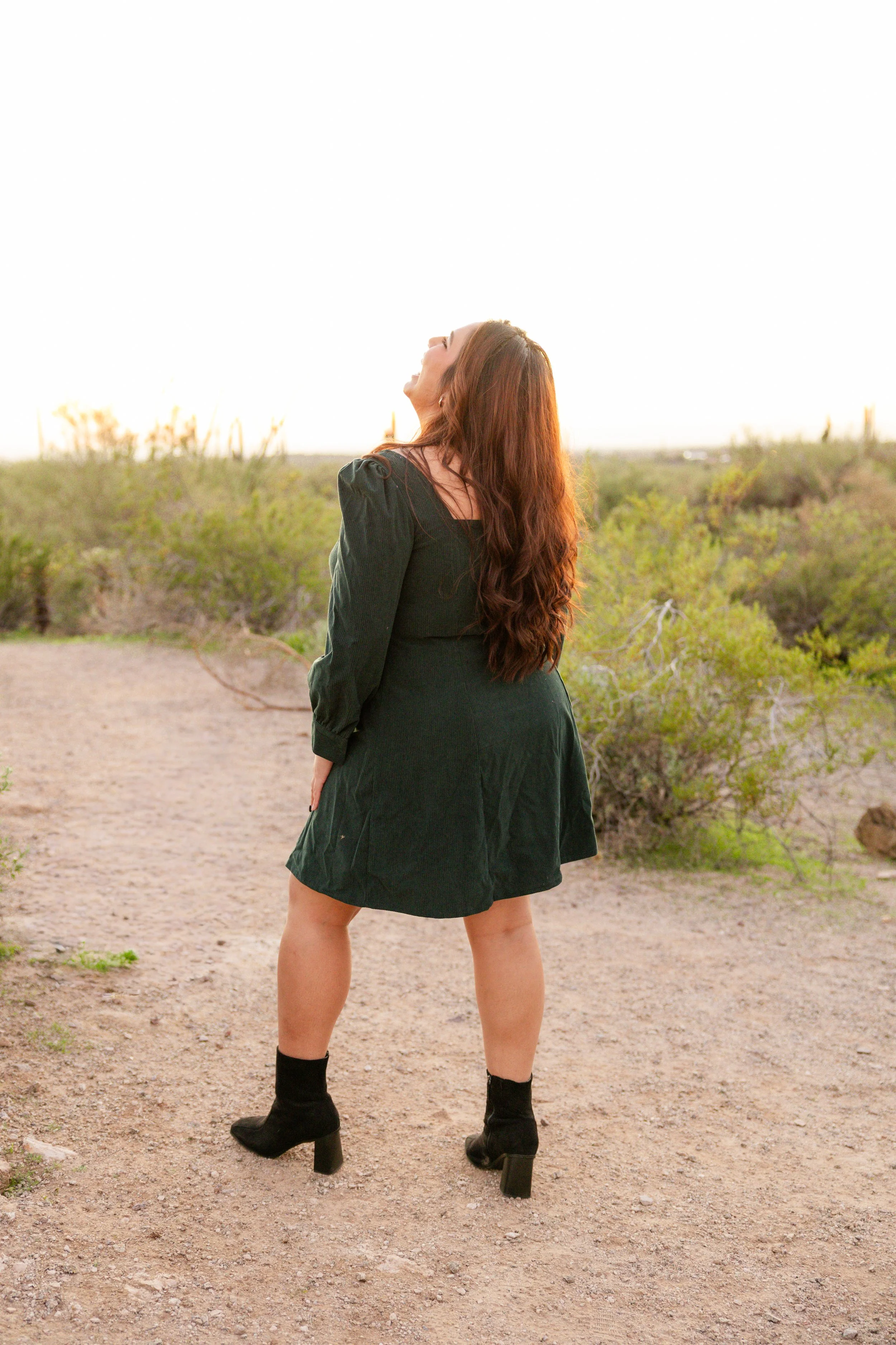 Woman in a black dress and high-heeled boots standing on dirt path in desert, laughing with head tilted back at sunset.