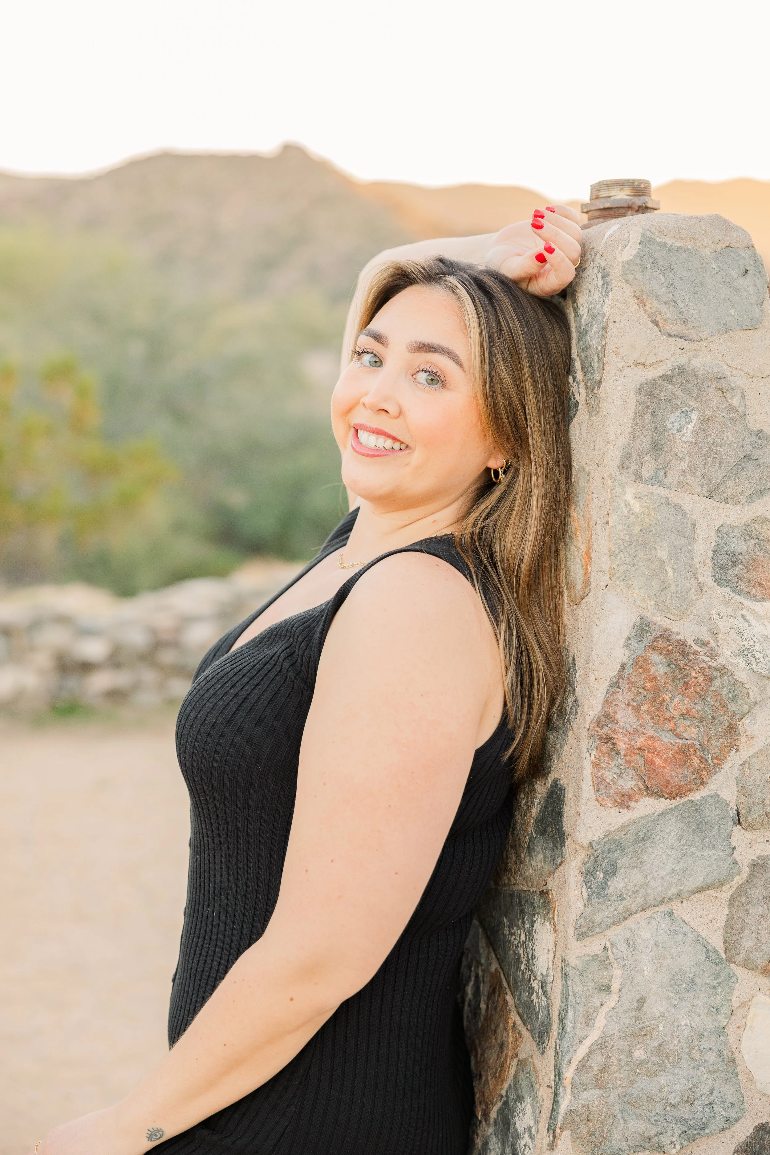 A woman leaning against a stone column outdoors during the daytime, smiling and looking at the camera. She has shoulder-length brown hair, green eyes, and is wearing a black sleeveless top with red nail polish.