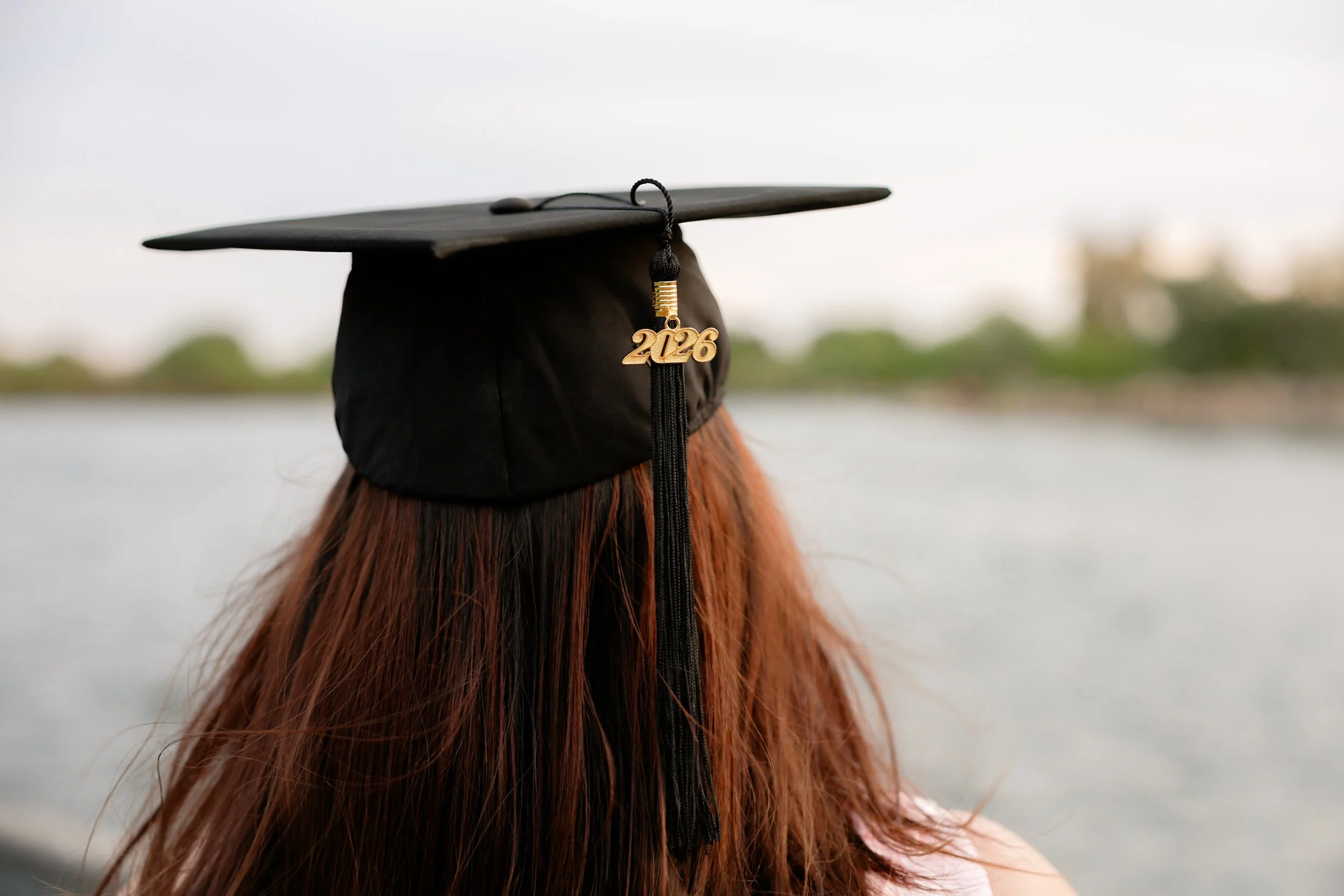 Person wearing a graduation cap with a gold charm reading '2026' on the tassel, outdoors near water with trees in the background.