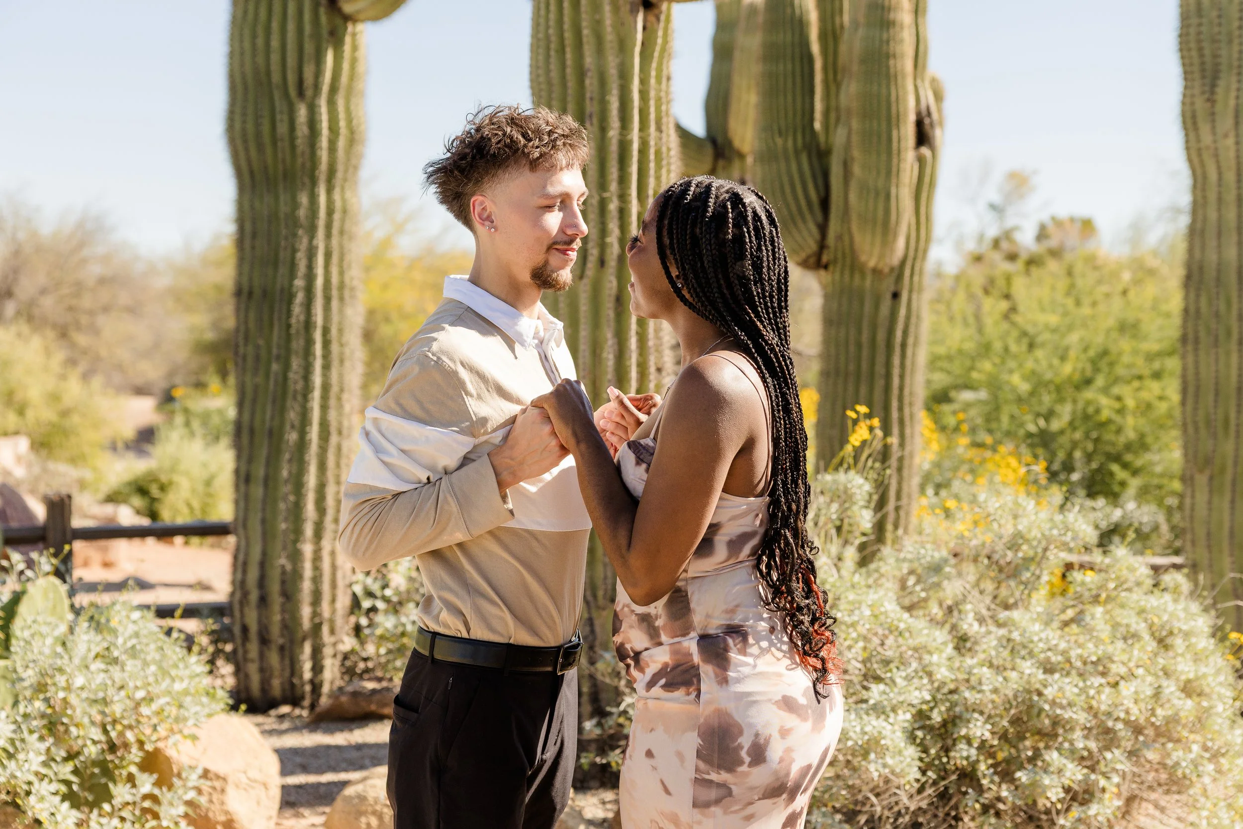 A couple holding hands, looking into each other's eyes in front of saguaro cacti with desert plants and a blue sky in the background.