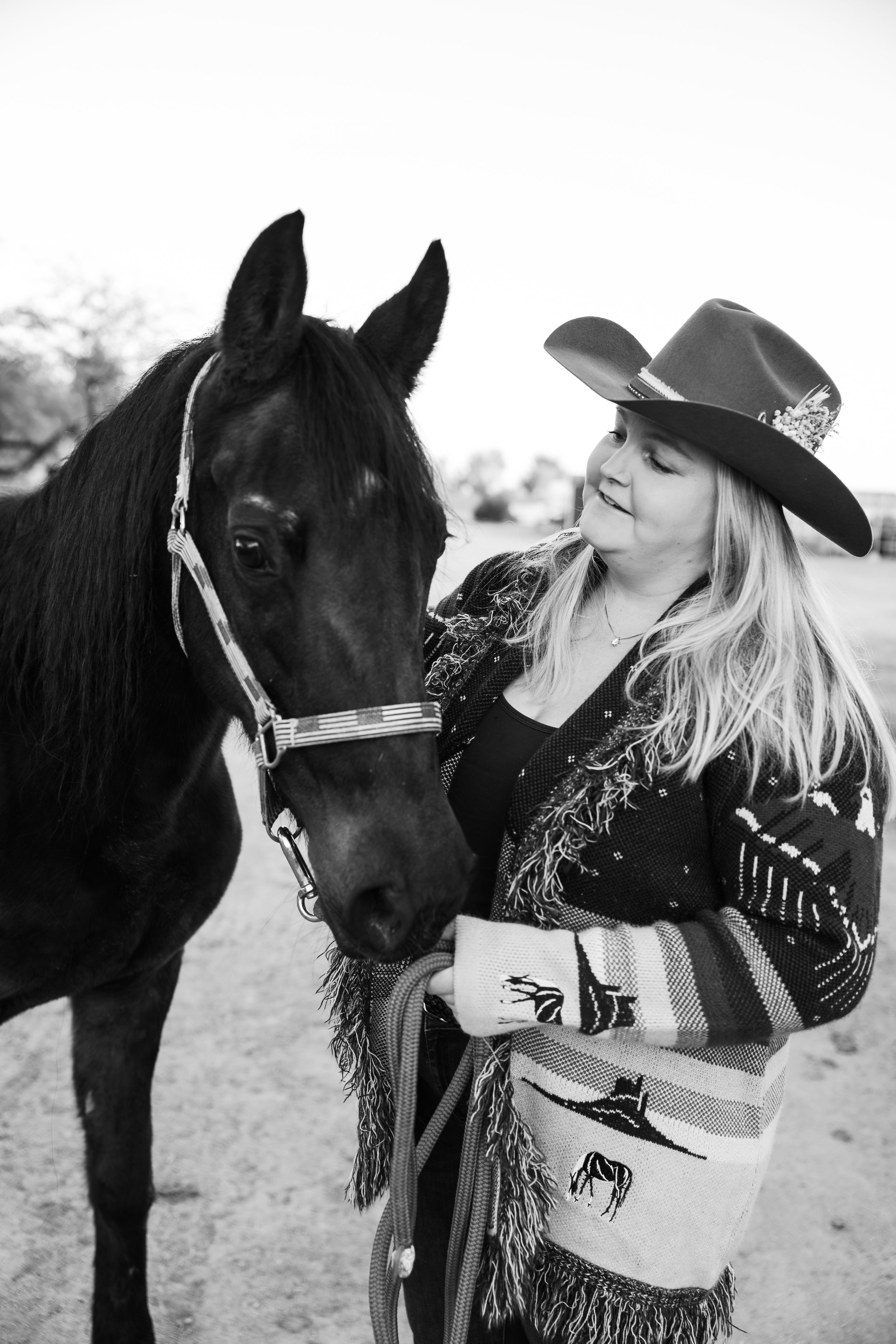 A girl with long blonde hair wearing a cowboy hat and a patterned sweater, standing outdoors and holding a horse's bridle. The horse is black and has a halter, and the girl is looking at the horse affectionately.