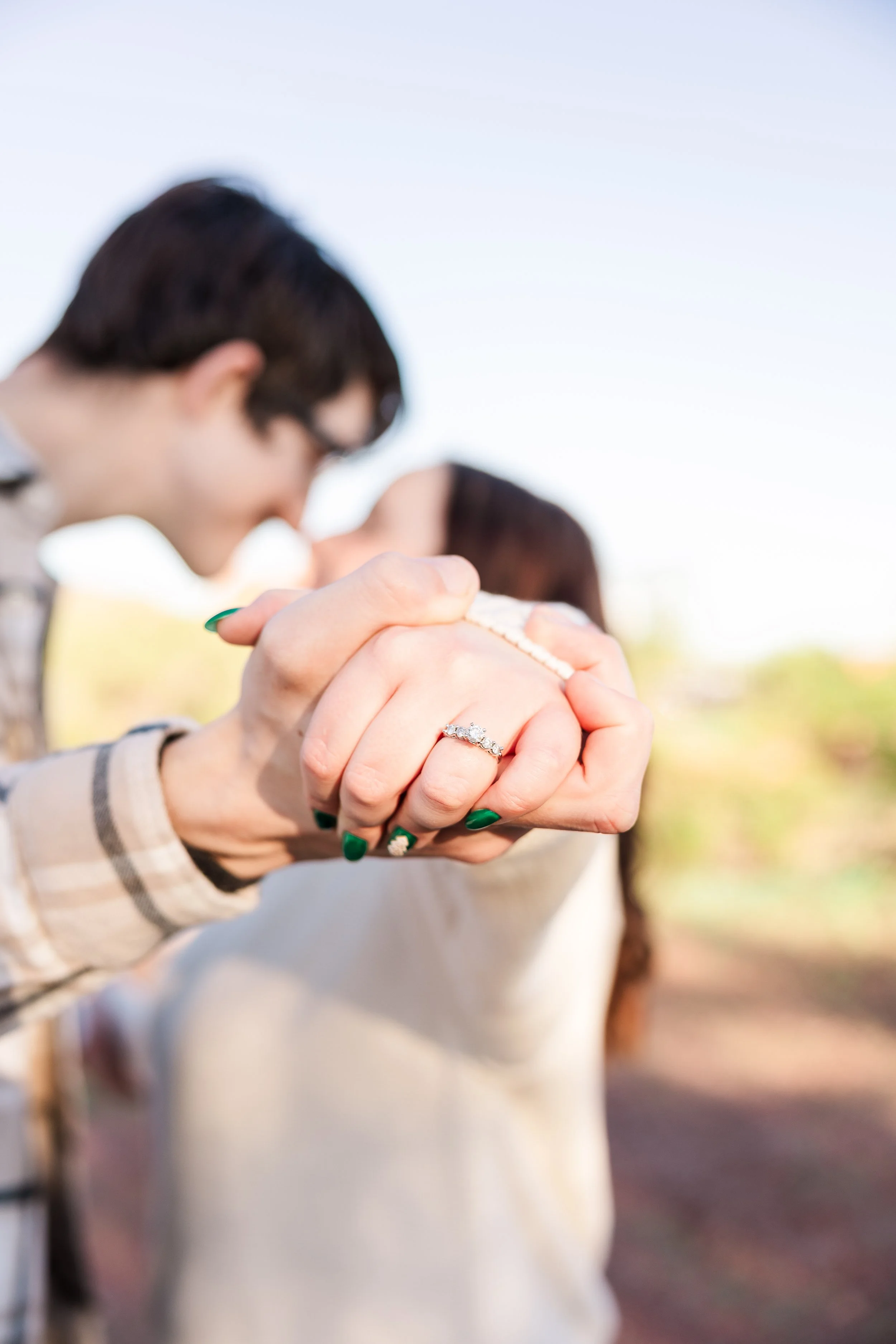 Close-up of a woman's hand with an engagement ring, holding a man’s hand in a kiss, with a blurry couple kissing in the background outdoors, under a clear sky.