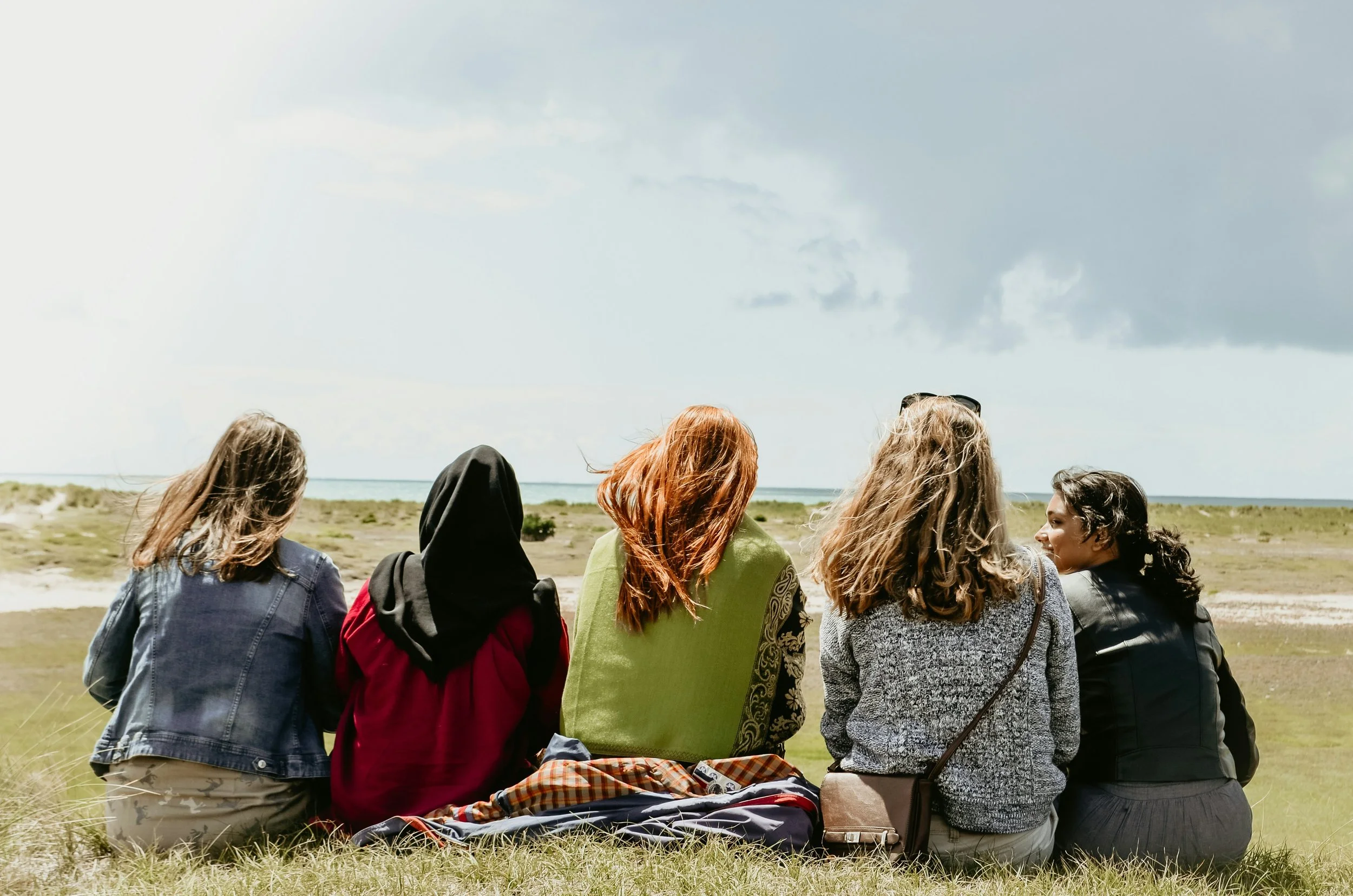 Five women sitting on grass, facing the ocean, with a clear sky above.