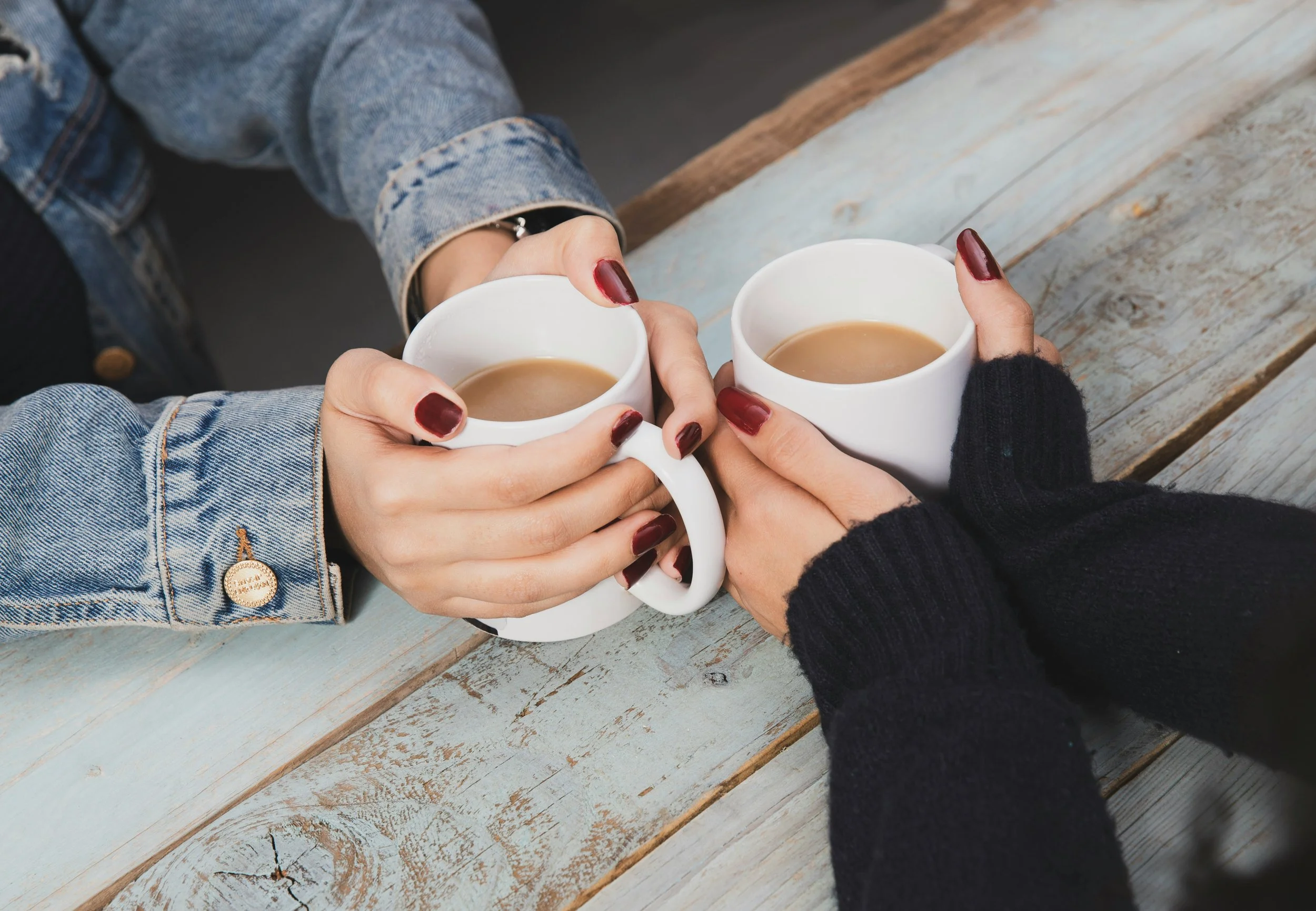 Two people holding white mugs of coffee on a wooden table, wearing denim and black sleeves.
