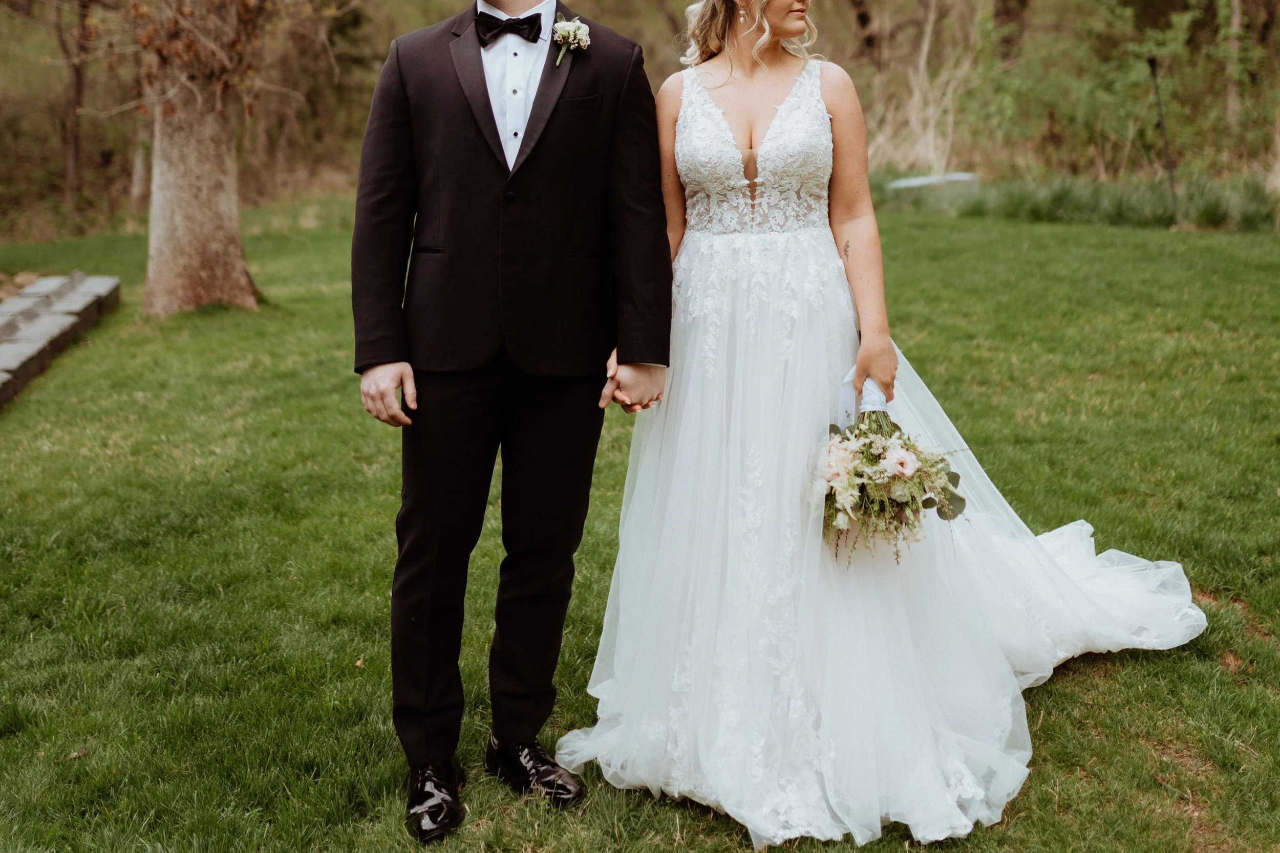 A bride and groom holding hands outdoors on a grassy area, with the bride in a white lace wedding gown holding a bouquet, and the groom in a black tuxedo with a bow tie.