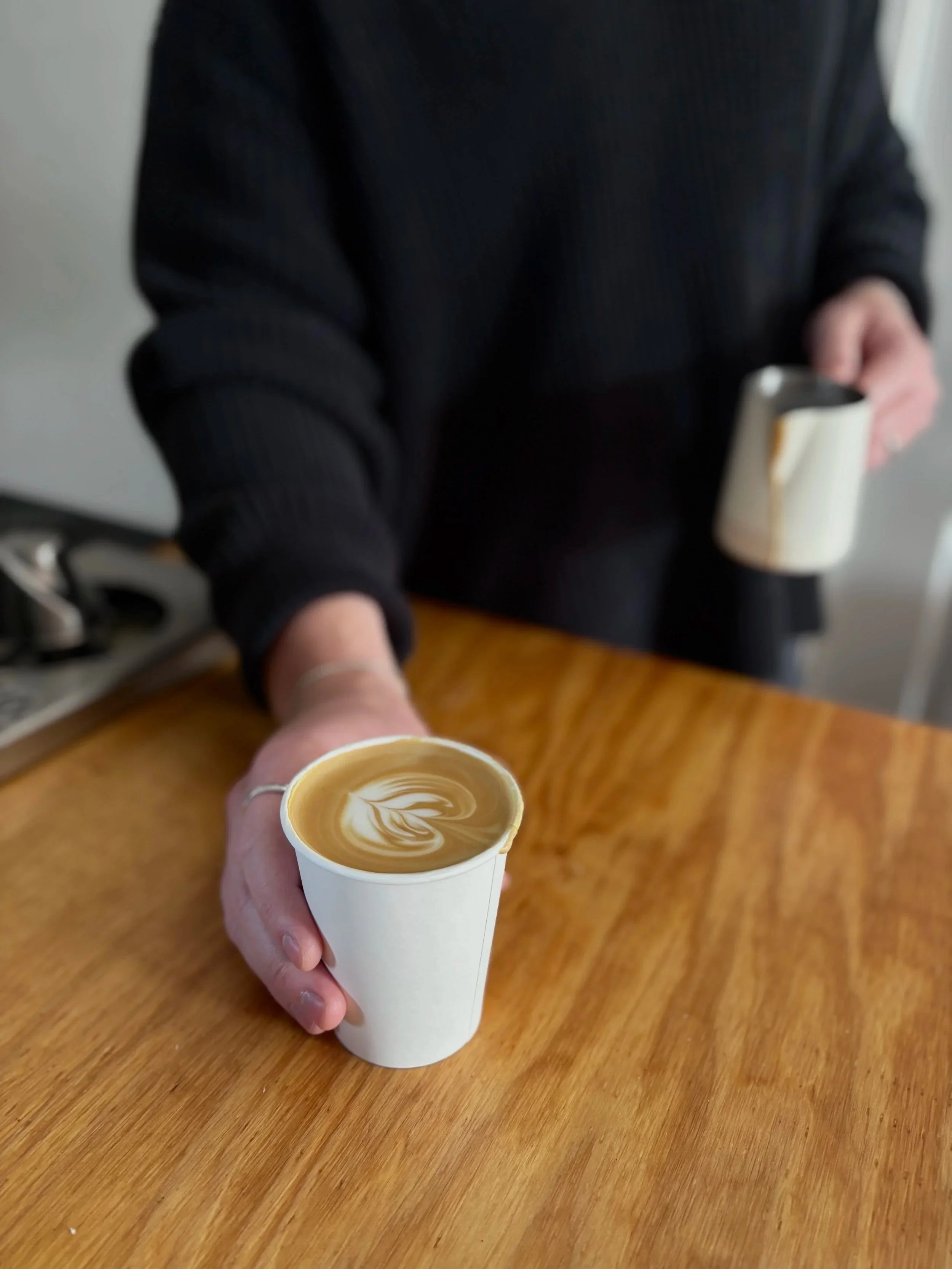 Eden & Oak Coffee barista holding a cup of coffee with latte art, offering it to the camera, with another cup in the background on a wooden table.