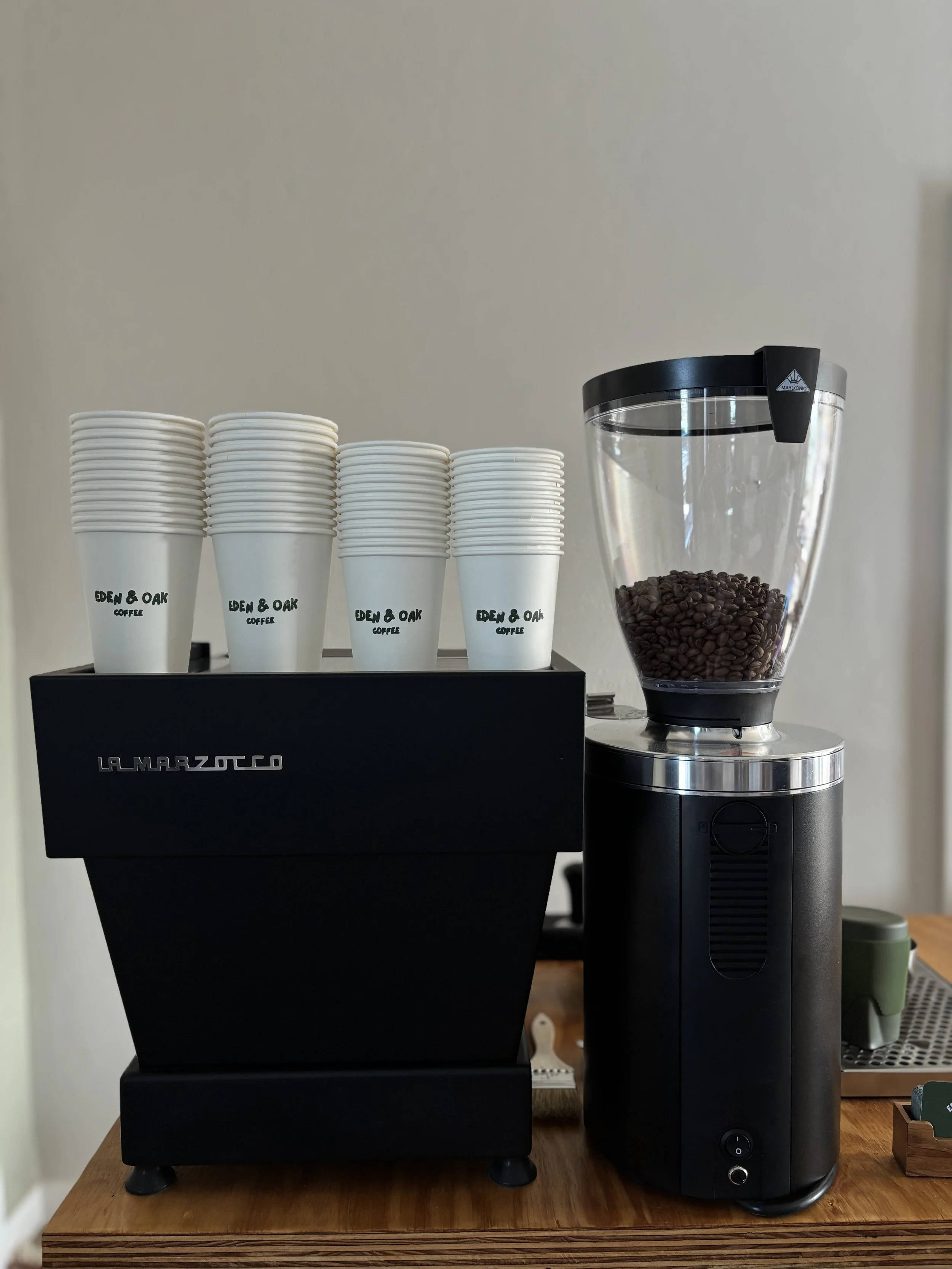Eden & Oak's coffee cart setup with a black La Marzocco espresso machine, a row of white cups labeled 'Eden & Oak Coffee, and a Mahlkonig espresso grinder filled with coffee beans..