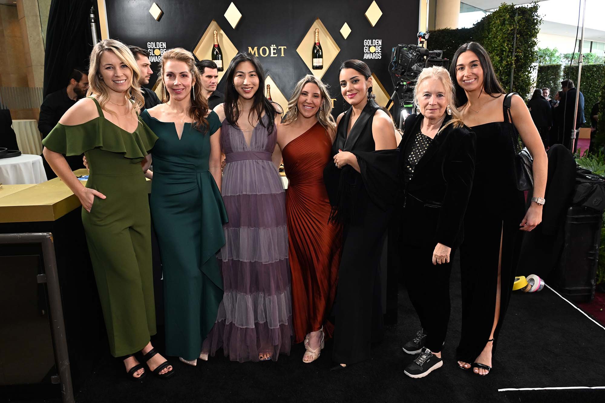 Group of eight women at a formal event, posing on a black carpet with a backdrop featuring Moët branding and champagne bottles. The women are dressed in elegant evening gowns and black attire.