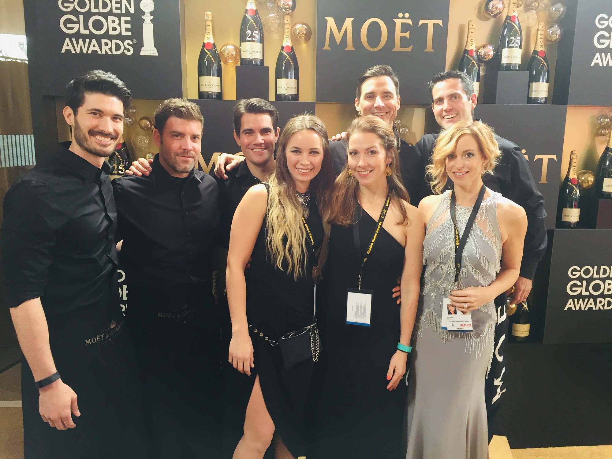 Group of eight people dressed in formal attire posing together at the Golden Globe Awards event, with a backdrop displaying the Moët branding and several champagne bottles.