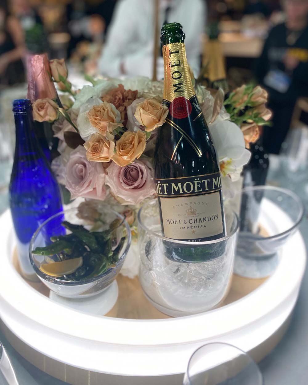 A bottle of Moët & Chandon Imperial champagne in ice surrounded by a floral arrangement of roses and other flowers on a glowing round table at a celebration or event.