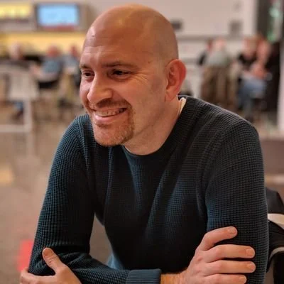 A smiling bald man in a dark sweater sitting at a table in a busy indoor setting.