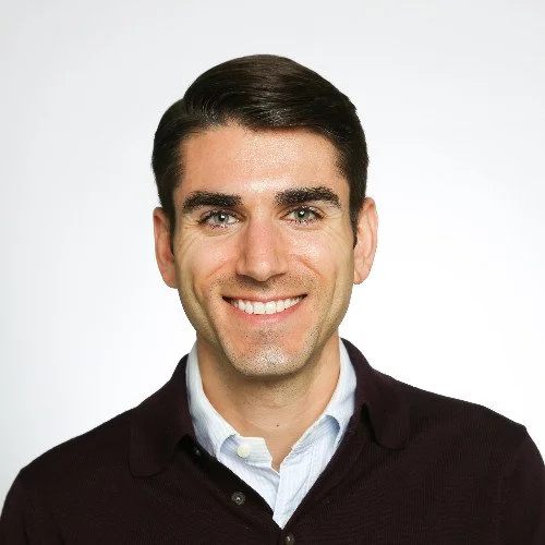 Headshot of a smiling man with dark hair, wearing a white shirt and a dark sweater, against a plain white background.