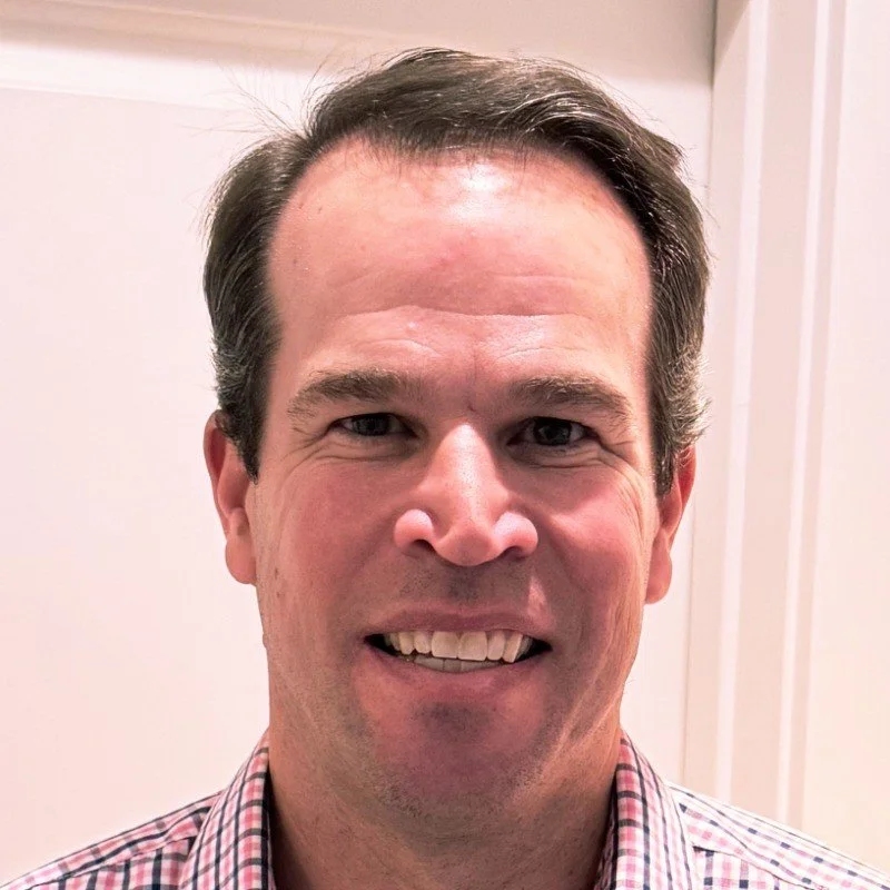 A smiling man with dark hair, wearing a checkered shirt, standing indoors in front of a beige wall.