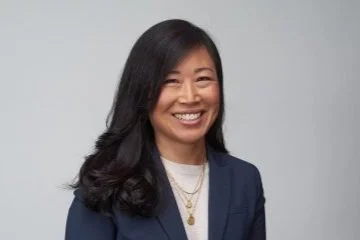 Portrait of a woman with long dark hair, wearing a navy blazer and gold necklaces, smiling against a gray background.
