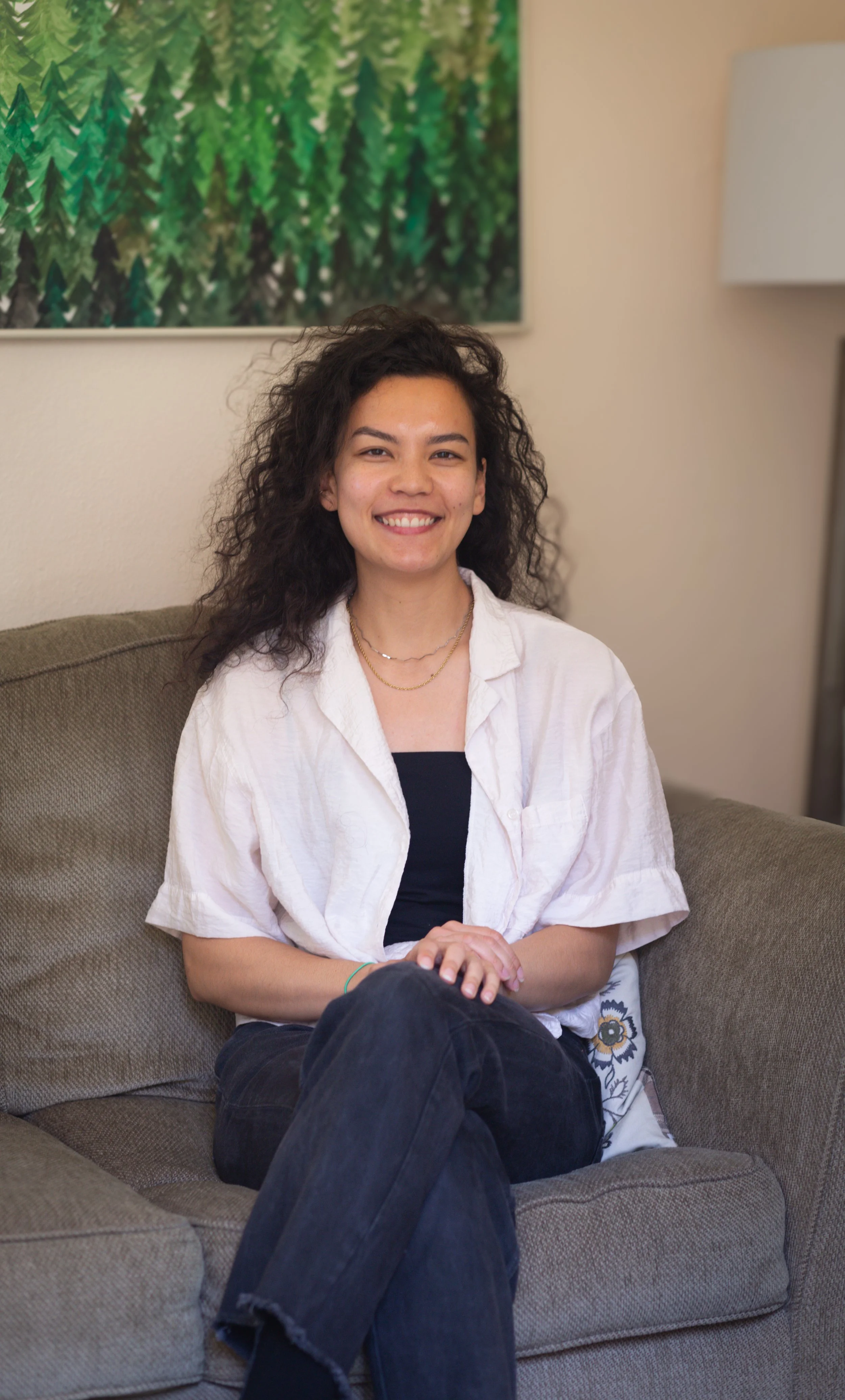 A young woman with curly dark hair sitting on a beige couch, wearing a white shirt over a black top, smiling at the camera in a cozy room with a forest-themed picture on the wall behind her.