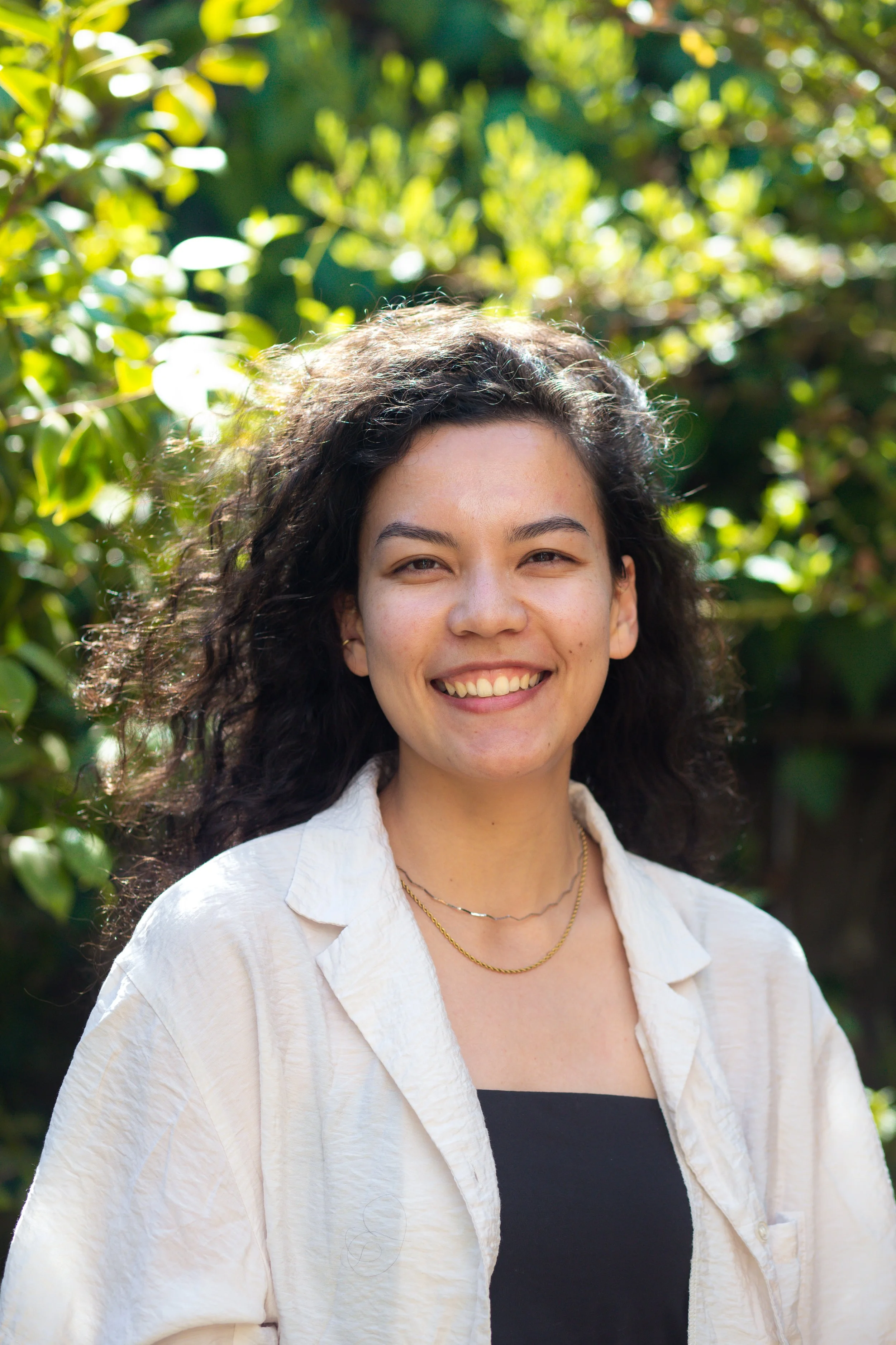 Smiling woman with curly dark hair wearing a beige shirt and layered necklaces outdoors with green foliage in the background.