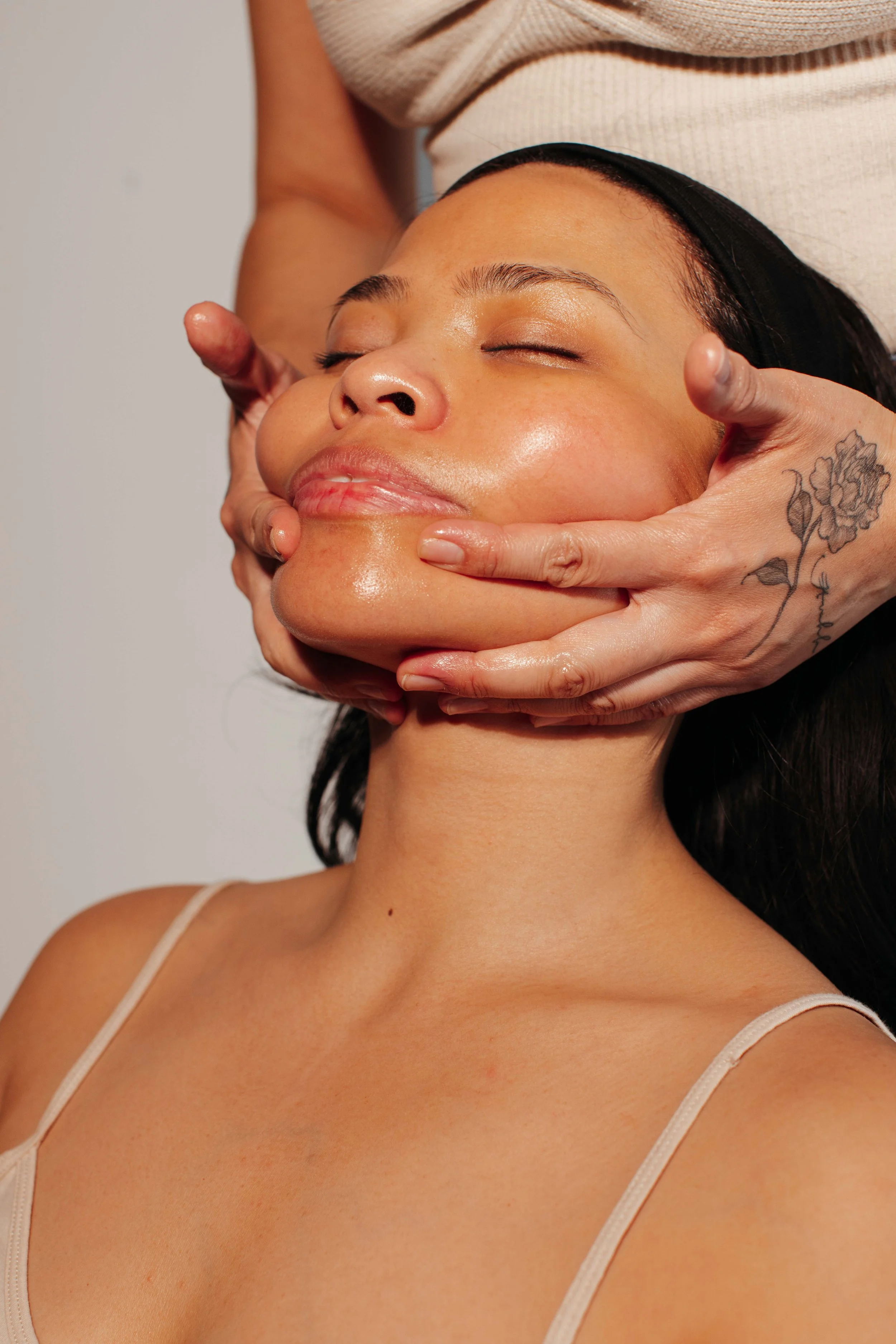 A woman receiving a facial massage with her eyes closed, a professional's hands gently holding her face, and a beige top visible.