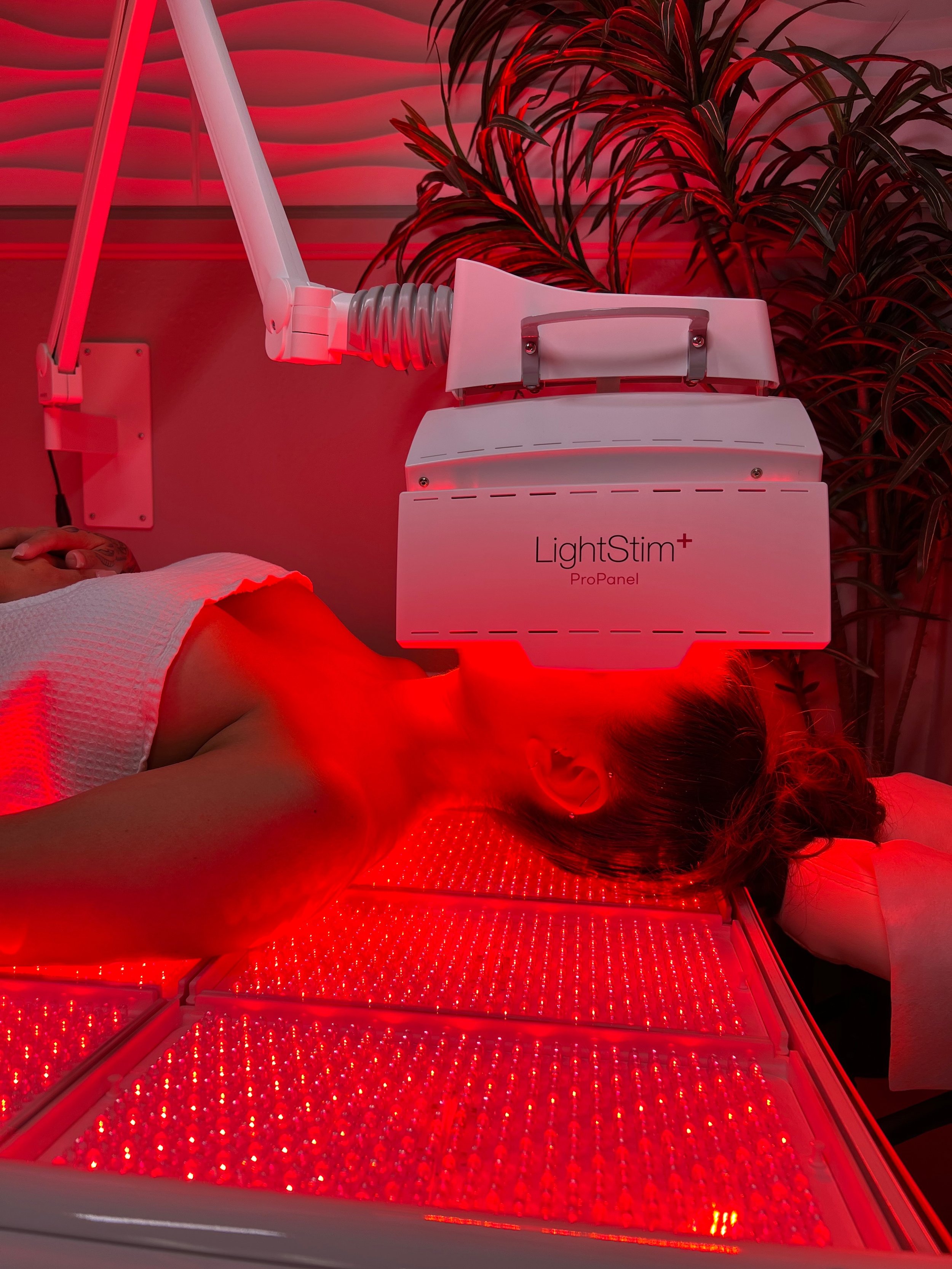 A person lying on a treatment bed receiving red light therapy with a LightStim ProPanel machine positioned above their chest, in a room with red lighting and a large plant in the background.