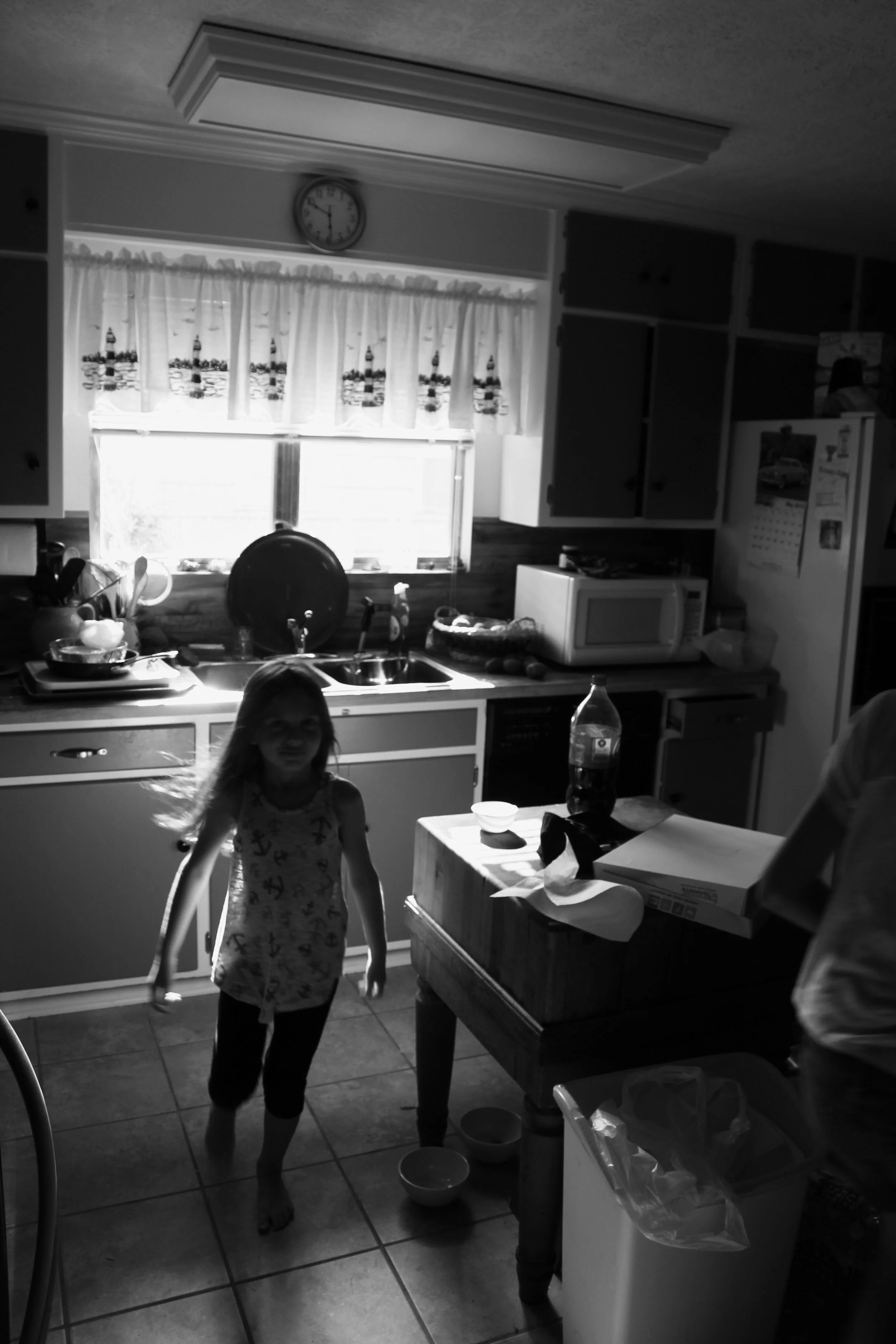 A black and white photo of a kitchen with a young girl walking toward the camera. The kitchen has a window with curtains, a sink, and various kitchen appliances on the counters. There is a table with a soda bottle, bowls, and a pizza box. Part of a p