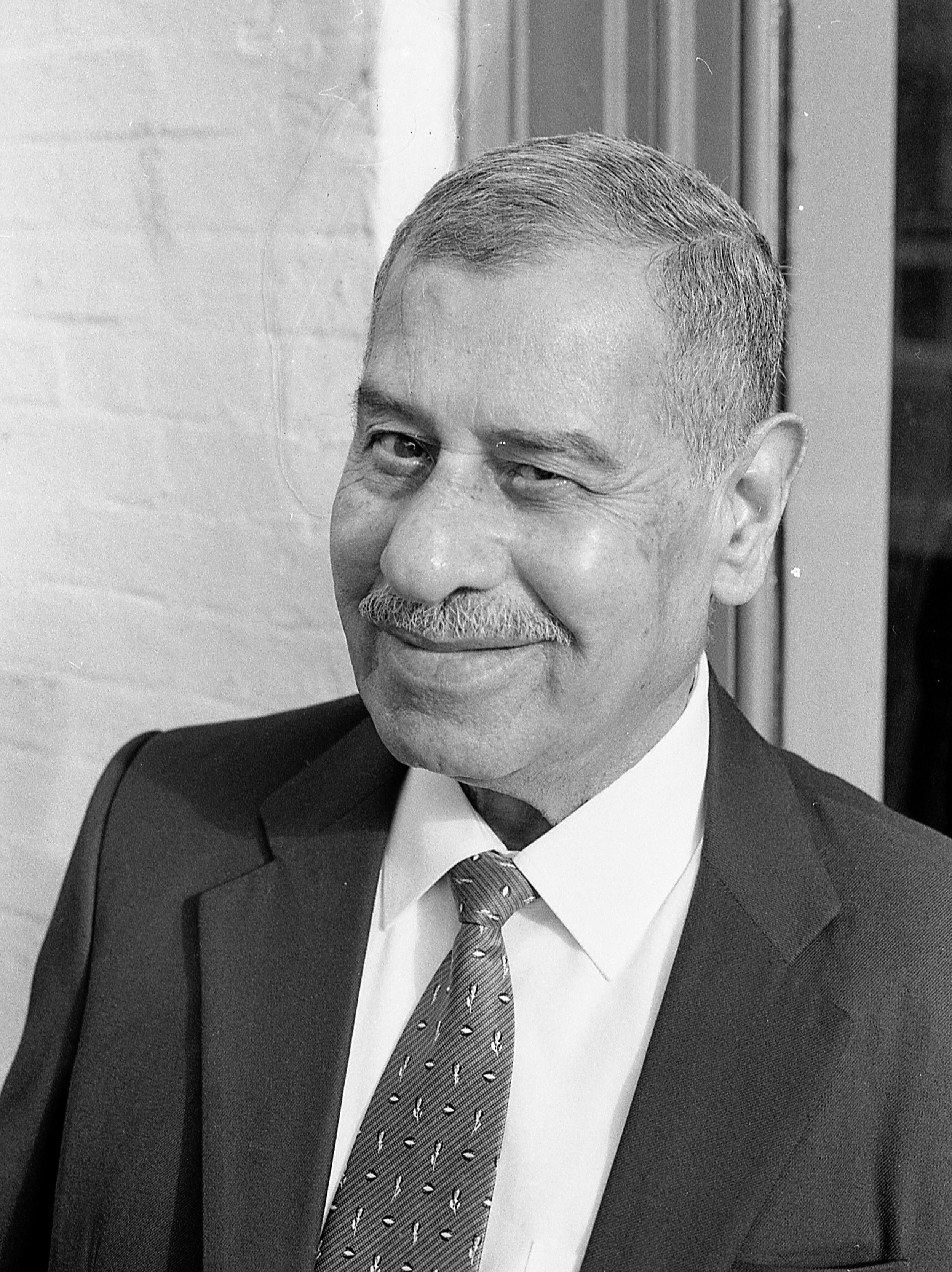 Black and white photo of a middle-aged man with a mustache, wearing a suit, white shirt, and patterned tie, smiling slightly while looking at the camera.
