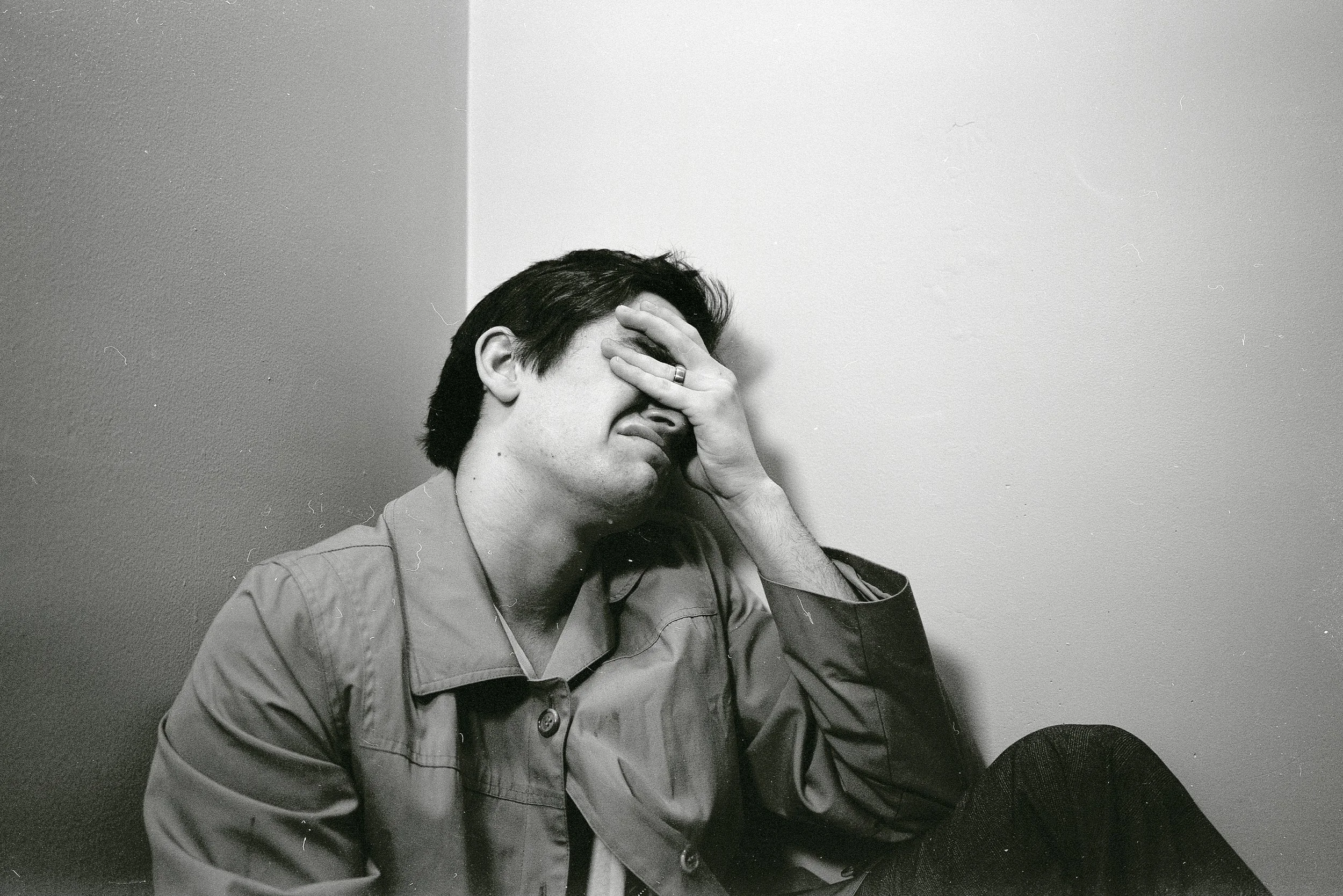 A man in a jacket sitting against a wall, with his hand covering part of his face, appearing distressed or upset.