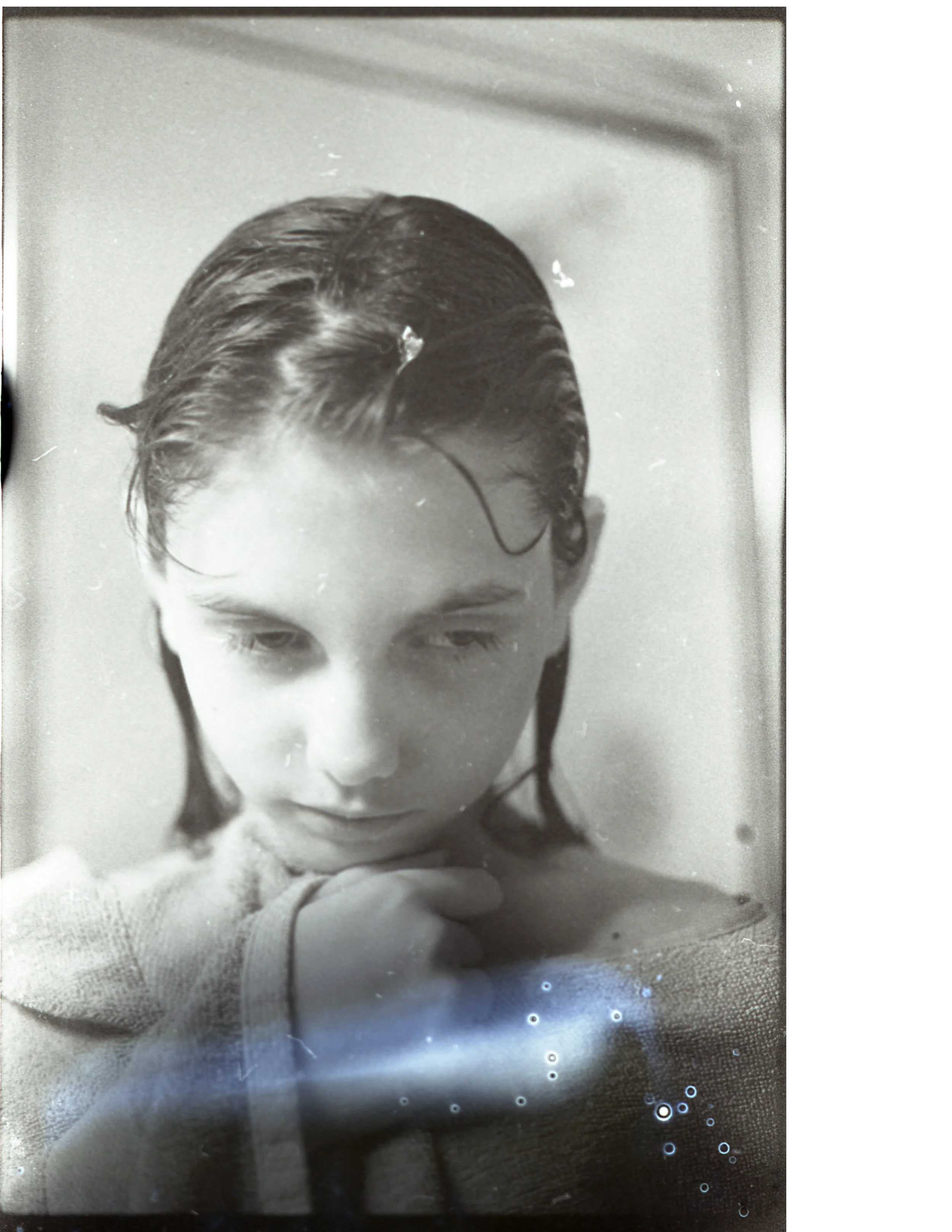 A black and white photo of a young woman with wet, slicked-back hair, looking down with her hand resting near her face.