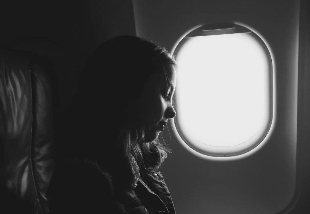 A woman with closed eyes sitting in an airplane seat next to a window, illuminated by natural light.