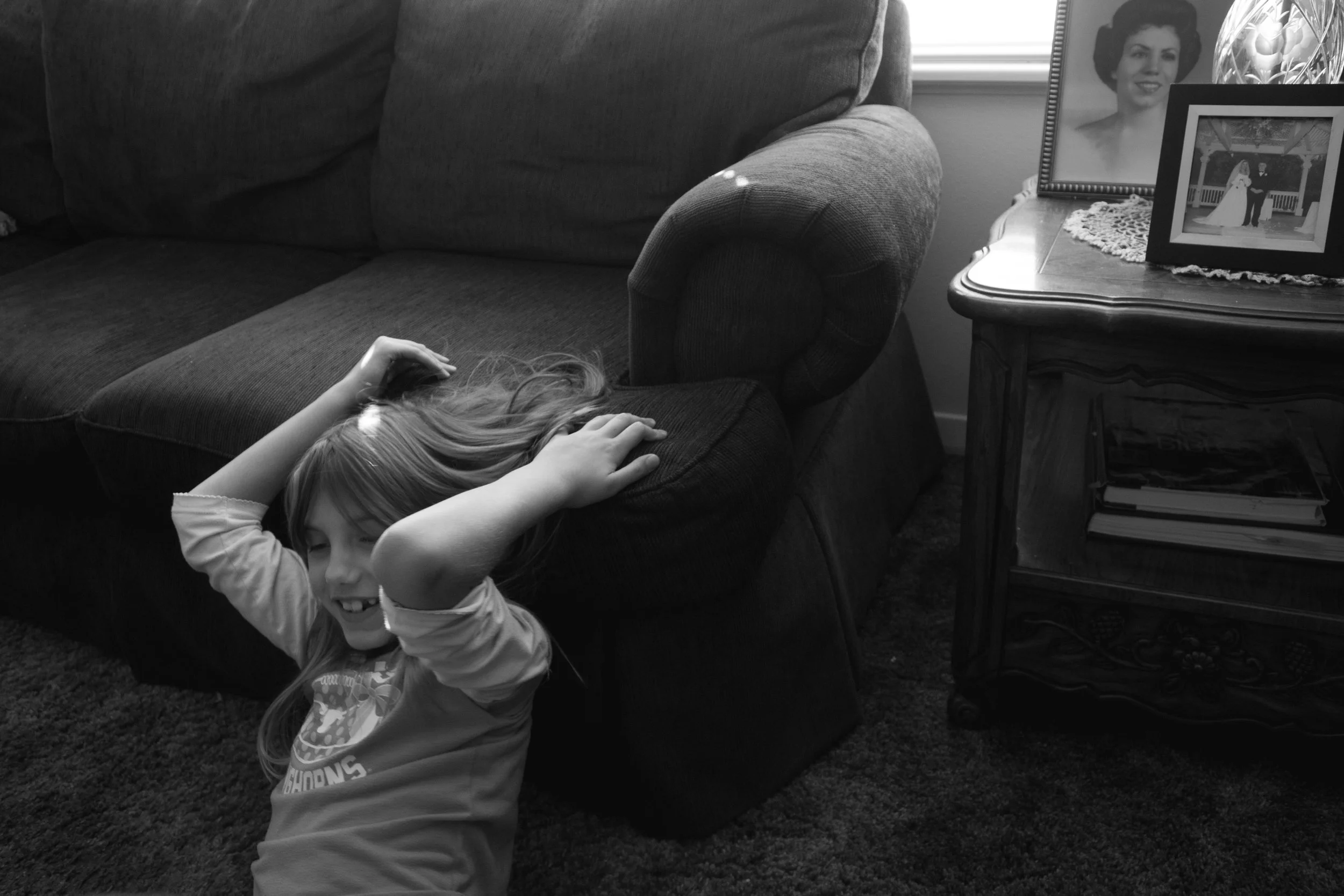 A young girl with long hair is sitting on the floor, leaning against a dark-colored sofa, smiling with her eyes closed. Her hands are on her head. Behind her, on a side table, are framed photographs, one of a woman smiling and another of a wedding sc