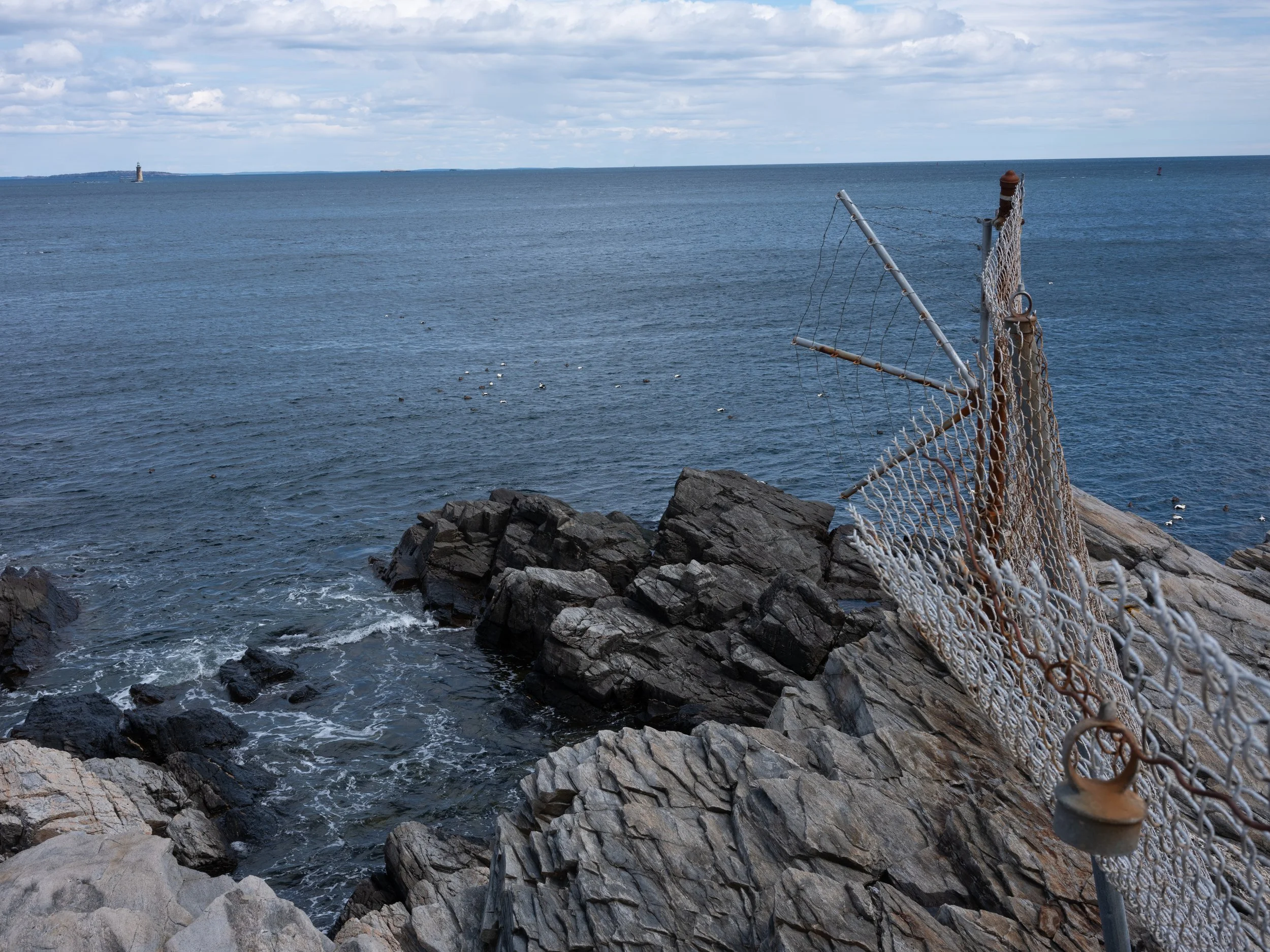 A rocky coast with a rusted chain-link fence and a view of the ocean extending to the horizon, with a lighthouse in the distance and seagulls floating on the water.